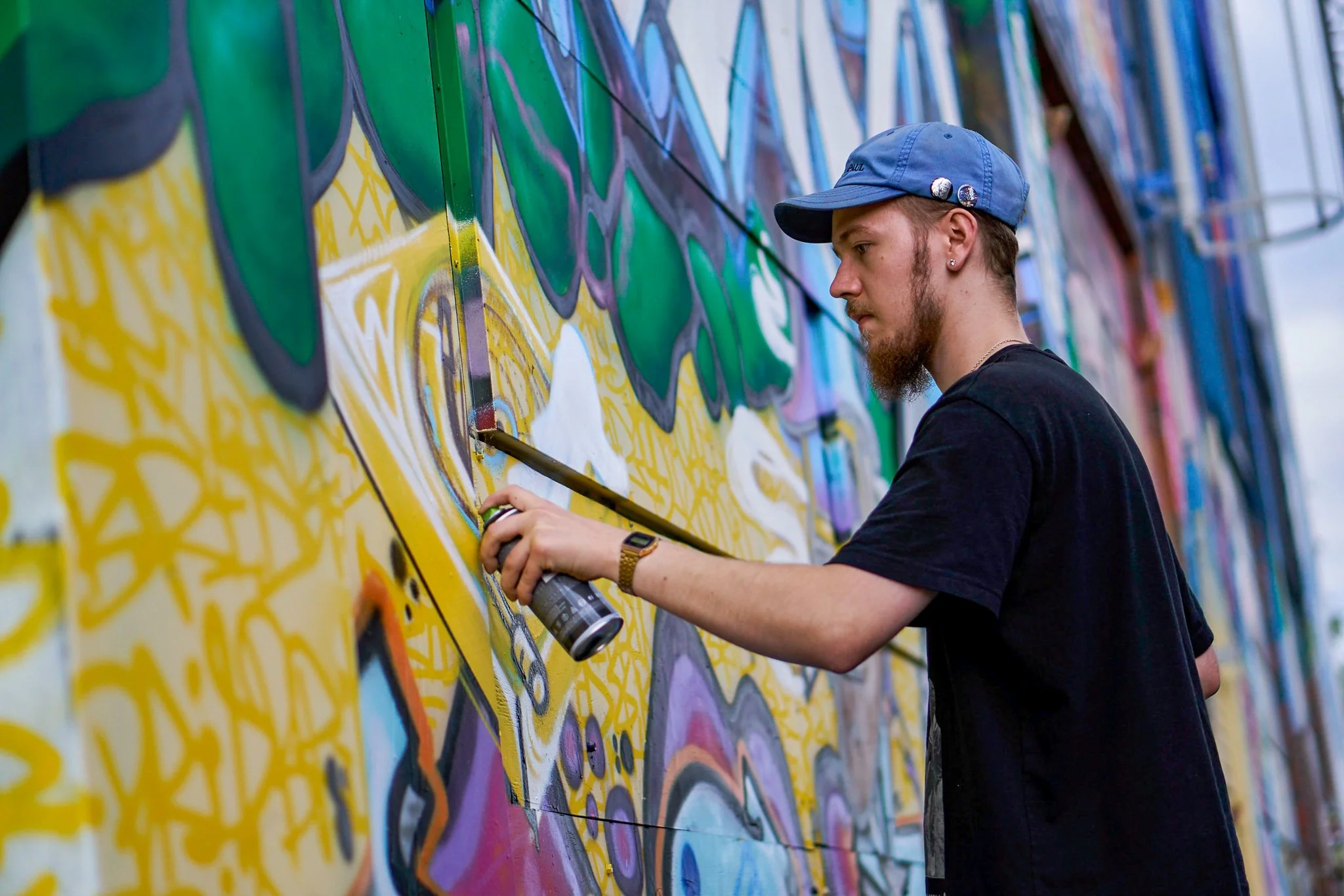 A young man with a beard, wearing a blue cap and black t-shirt, spray-painting graffiti on a colorful wall.