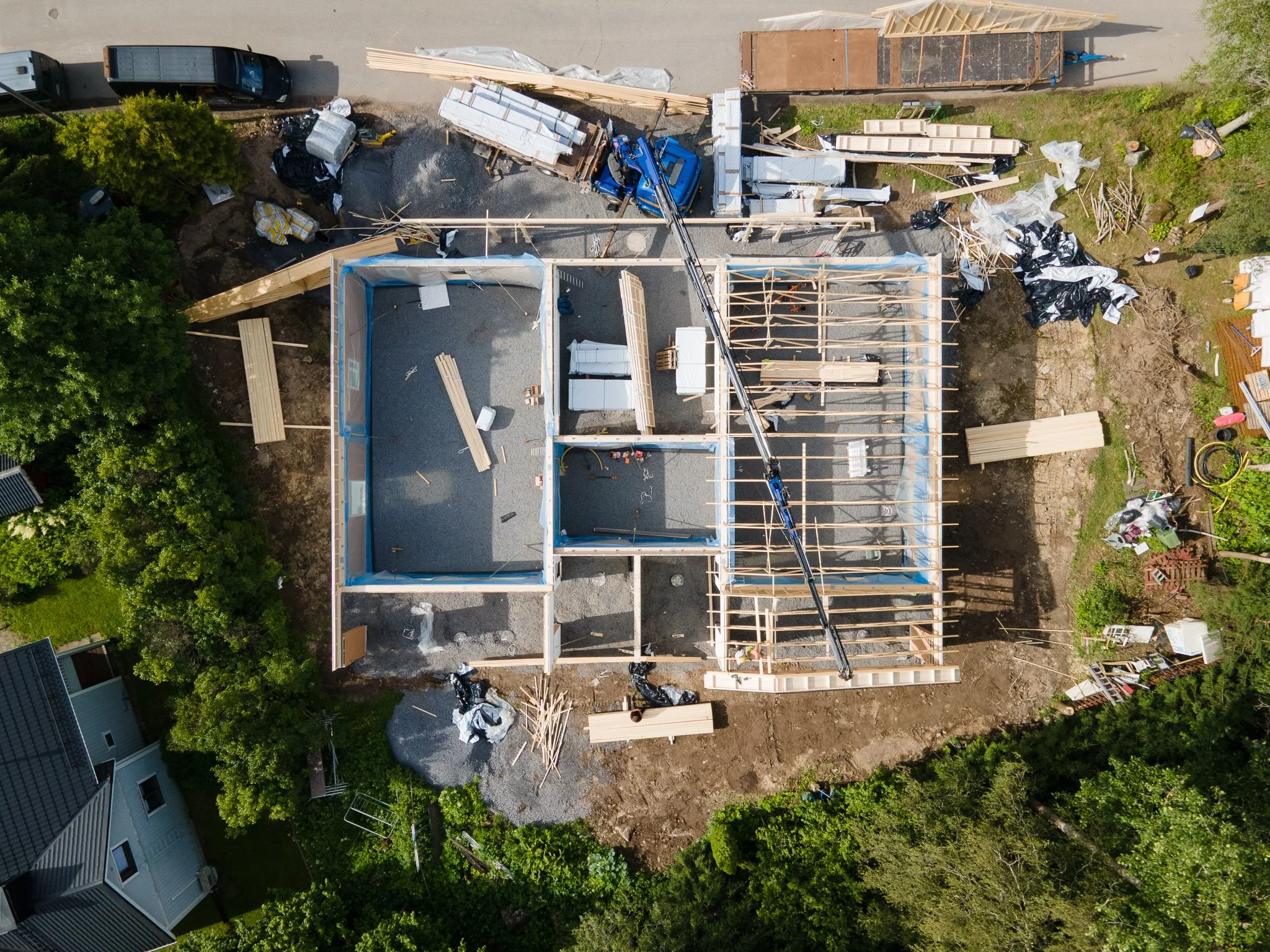 Aerial view of a house under construction with wooden framing and construction materials scattered around, surrounded by trees and neighboring houses.