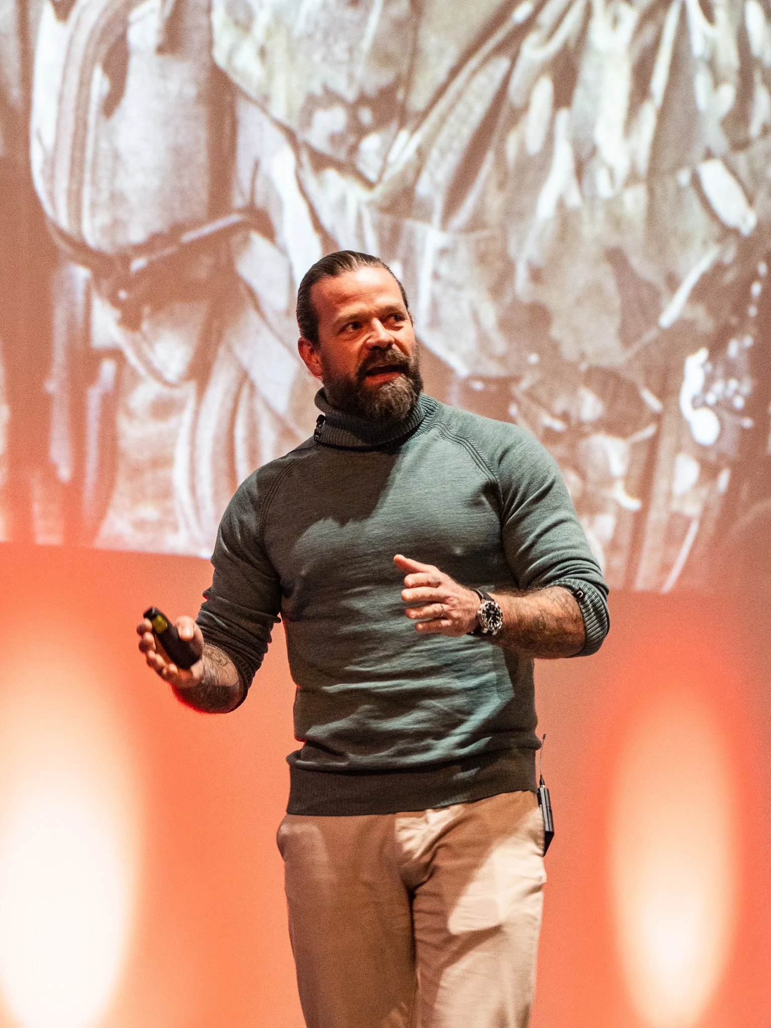 A man with a beard, wearing a gray sweater and beige pants, giving a presentation on stage with a large vintage map projected behind him.