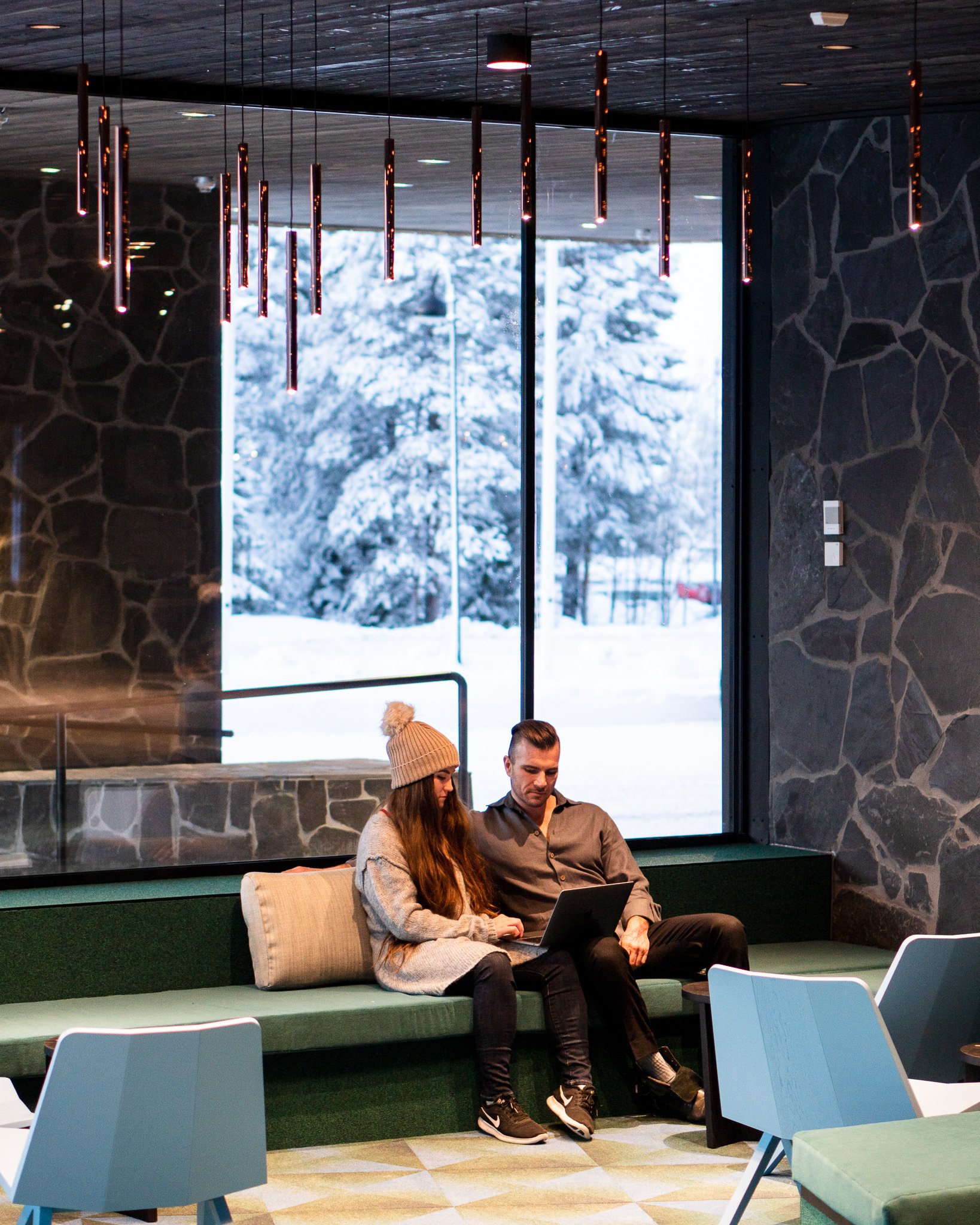 A young woman and man sit on a green couch in a modern indoor space near large window, looking at a laptop, with snow-covered trees seen outside.