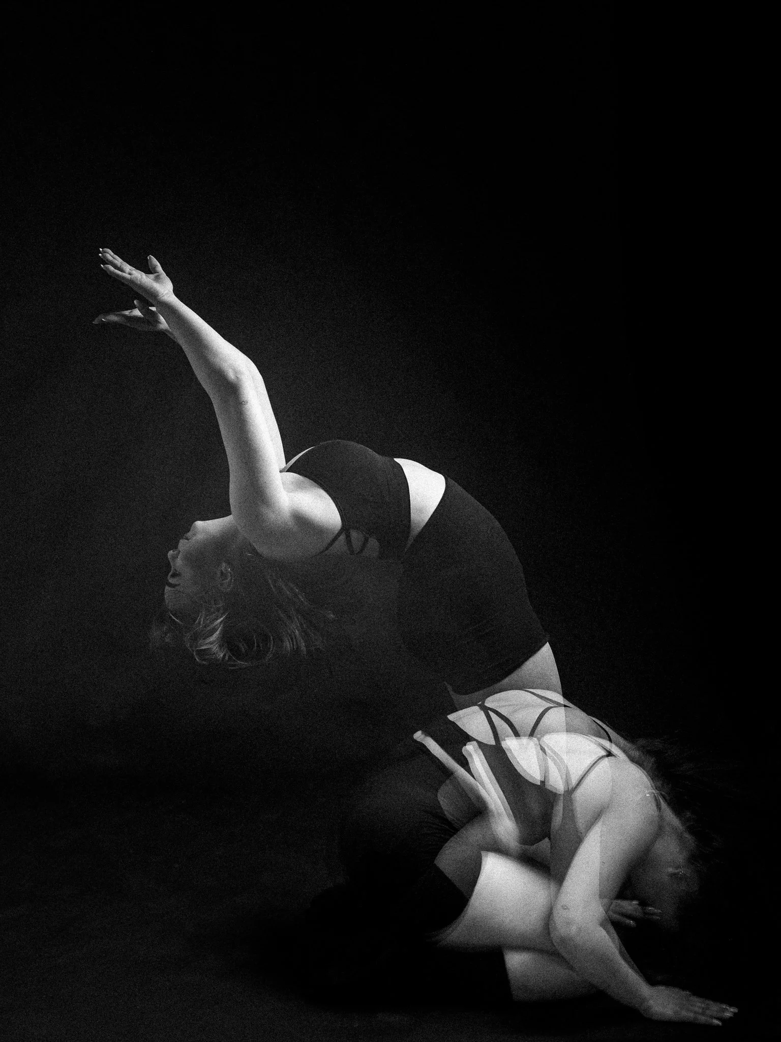 Two women practicing yoga on a dark background, one in a backbend pose and the other supporting with her hands and knees.