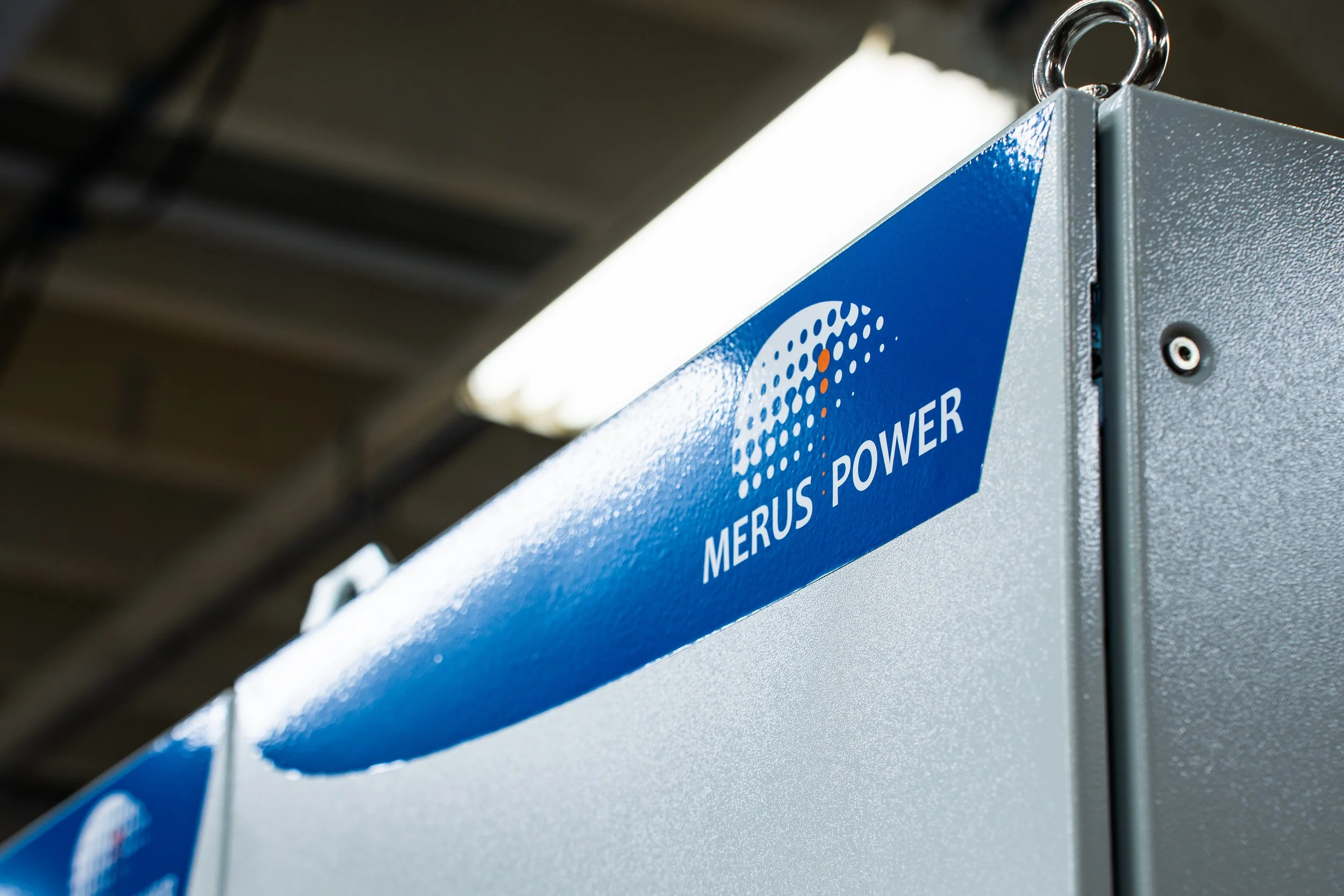 Close-up of a Merus Power electronic device with blue and gray casing, displaying the Merus Power logo, mounted on a wall or structure in an industrial setting.
