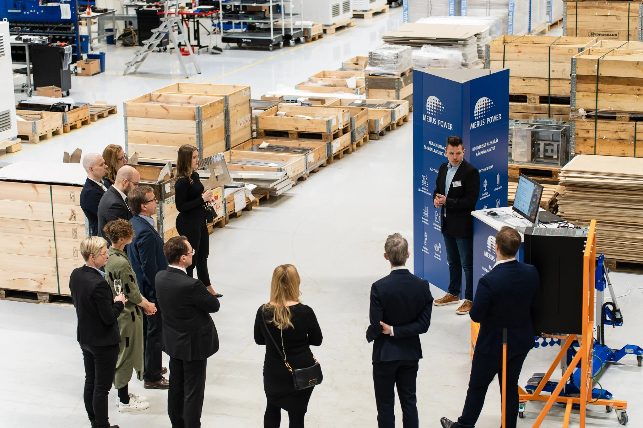 A presenter stands in front of a group of people inside a warehouse or factory setting. The audience is listening attentively, some holding glasses. The presenter is near a blue screen with the words 'Merus Power' and various icons, next to a compute