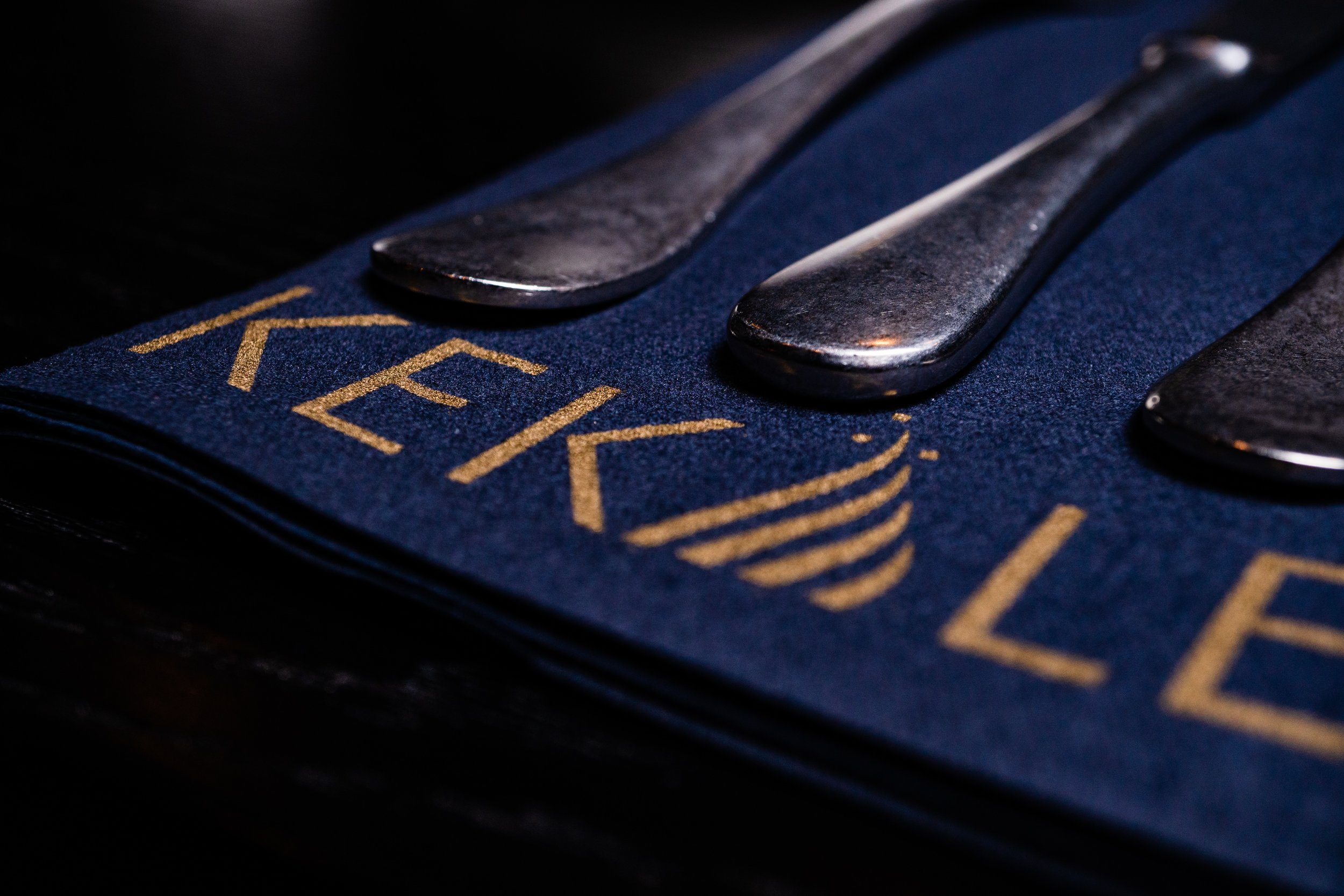 Close-up of metallic cutlery on a dark blue napkin with gold lettering, possibly part of a restaurant or event setting.