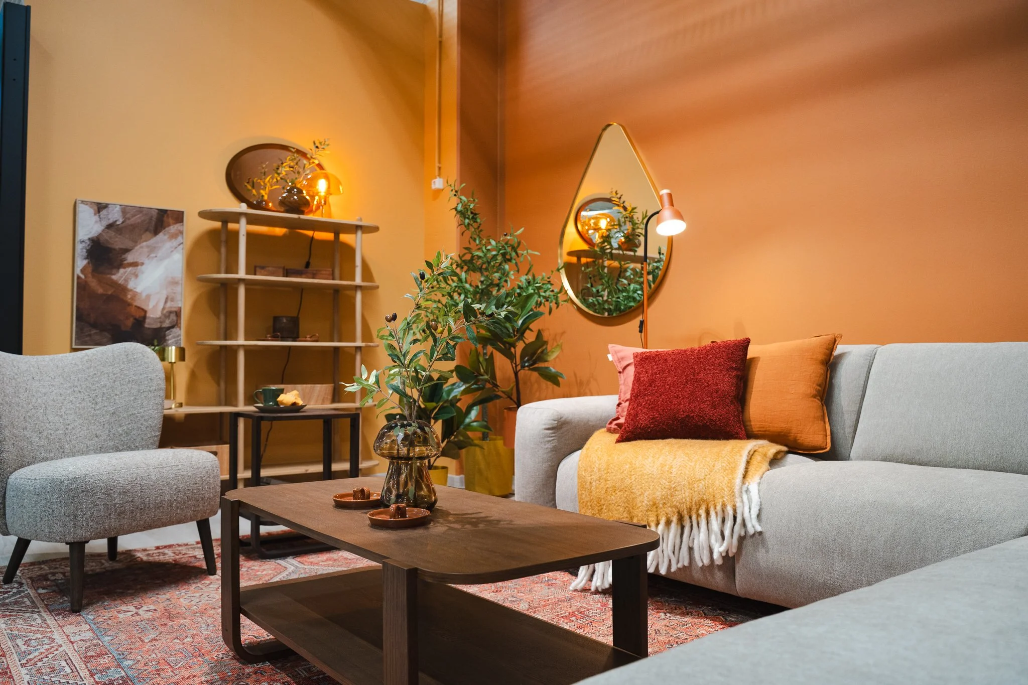Living room with beige sofa, gray armchair, wooden coffee table, colorful pillows, yellow blanket, potted plant, mirror, shelf, abstract art, warm lighting.