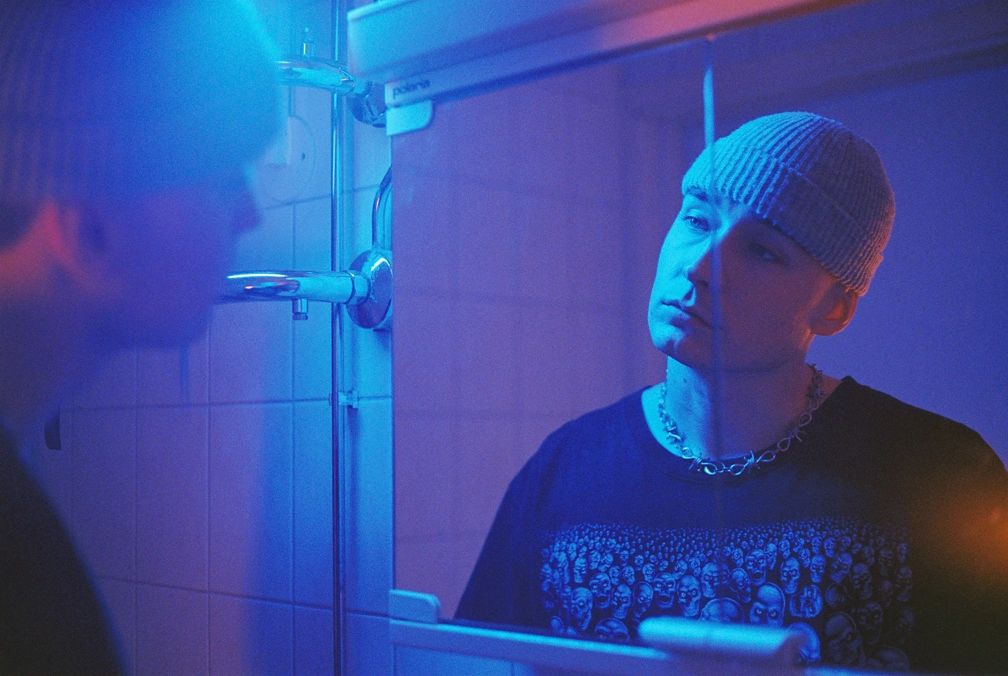 A young person wearing a knit cap and a chain necklace looking at his reflection in a mirror in a room lit with blue and purple lights.