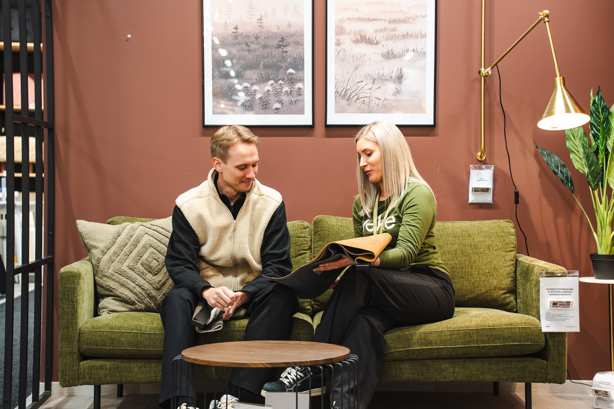A young man and woman sit on a green sofa in a furniture store, looking at a fabric sample or upholstery swatch together.