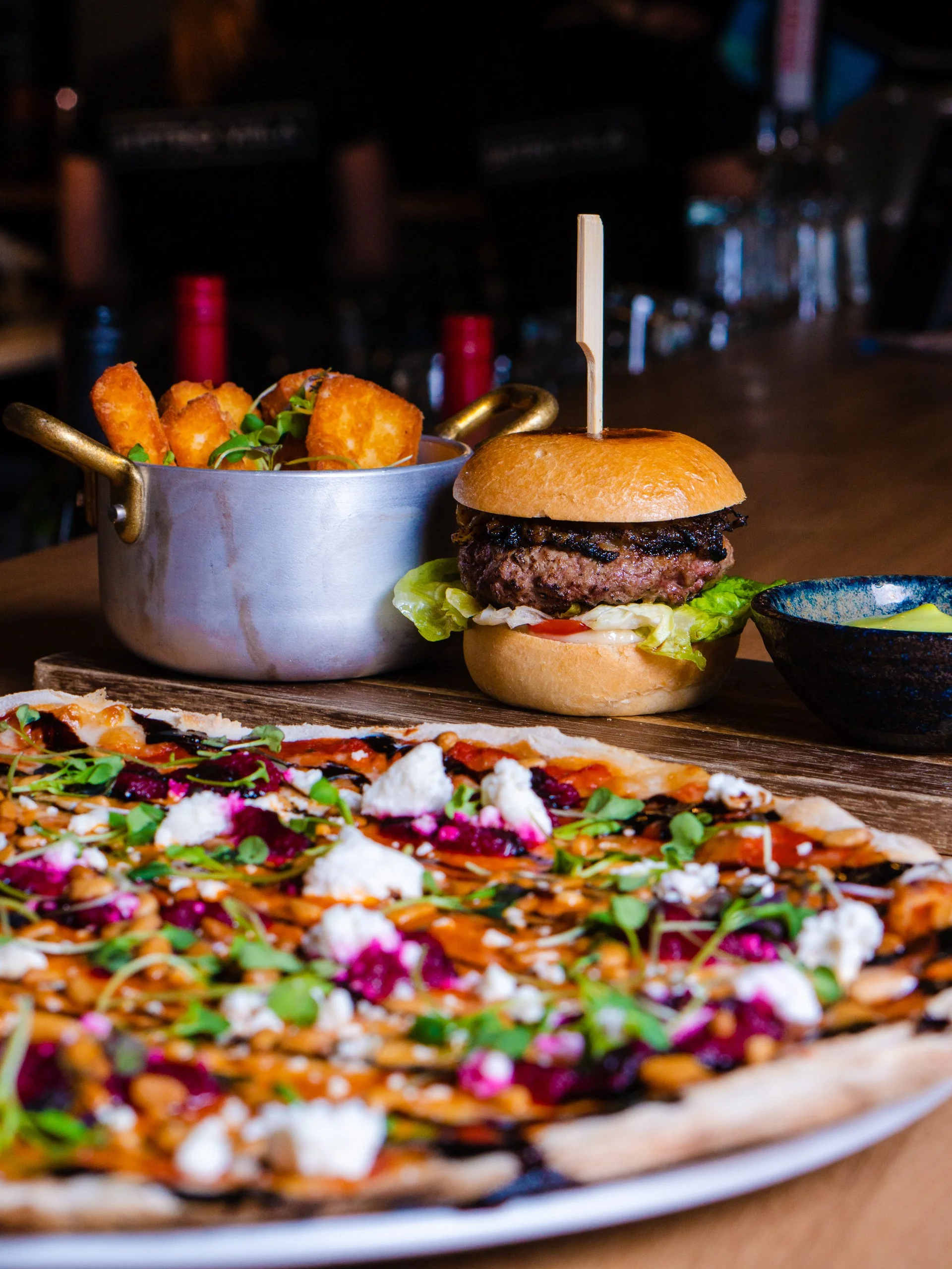 Close-up of a pizza with toppings, a burger with lettuce and tomato, and a side of tater tots with dipping sauce on a wooden table in a restaurant setting.