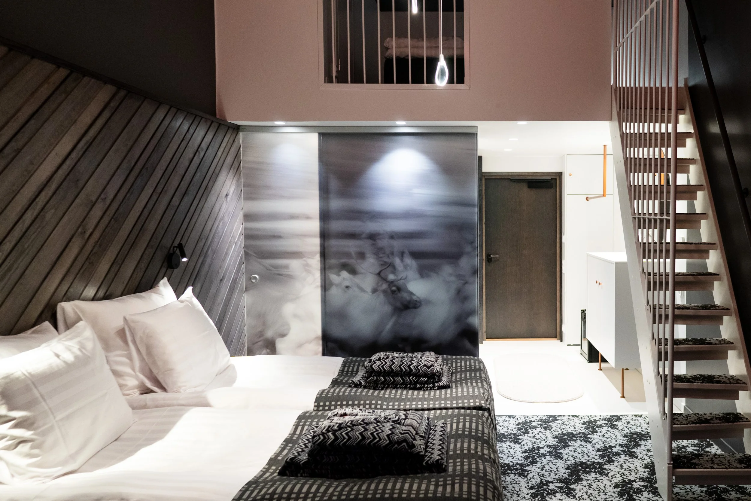 Modern hotel room with a bed, black and white patterned bedding, wooden wall panel, a wolf image on a sliding door, stairs, and various furniture.