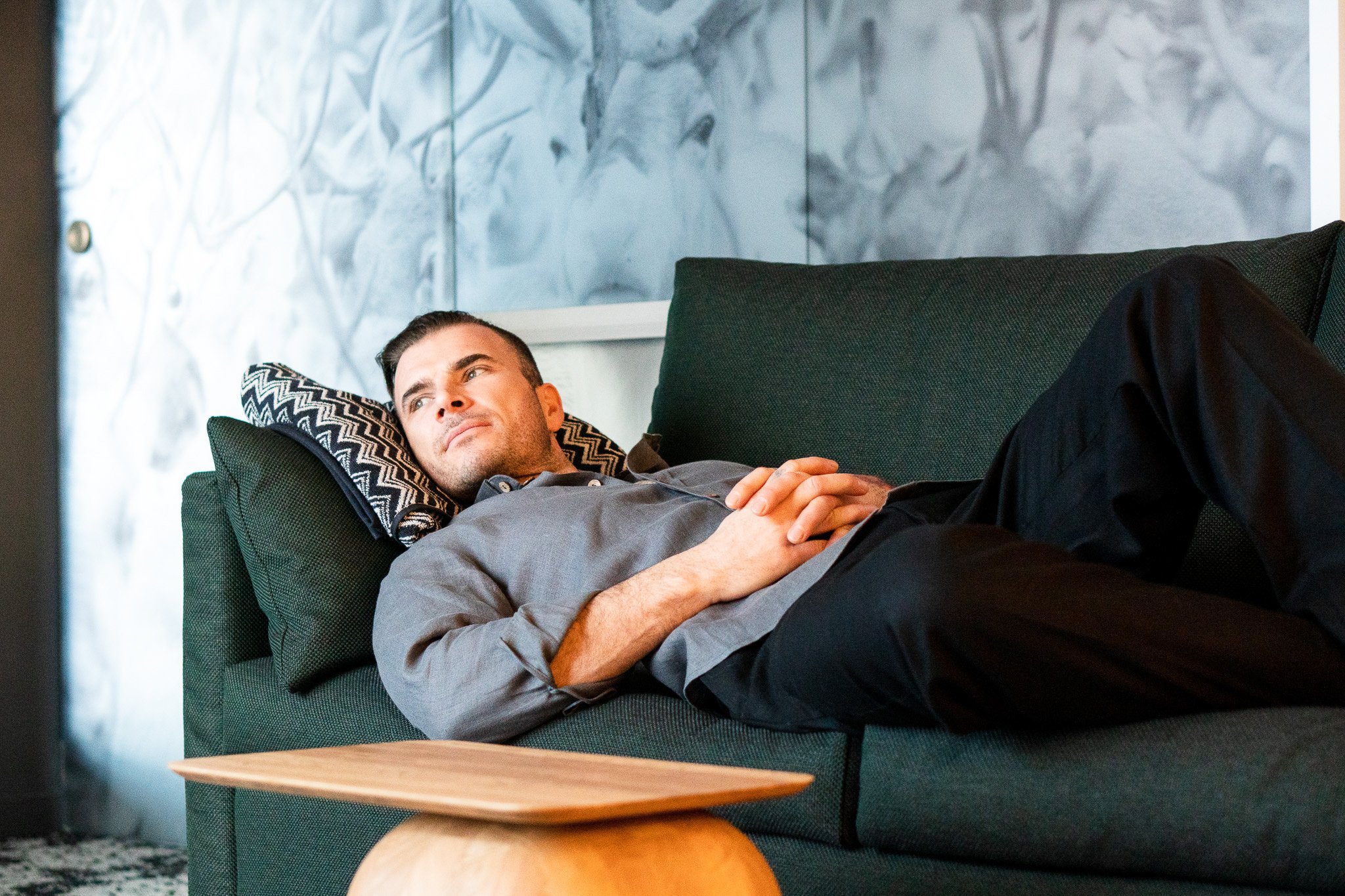 A man lying on a dark green couch with a pillow, looking thoughtfully into the distance with his hands clasped on his stomach, next to a wooden side table.
