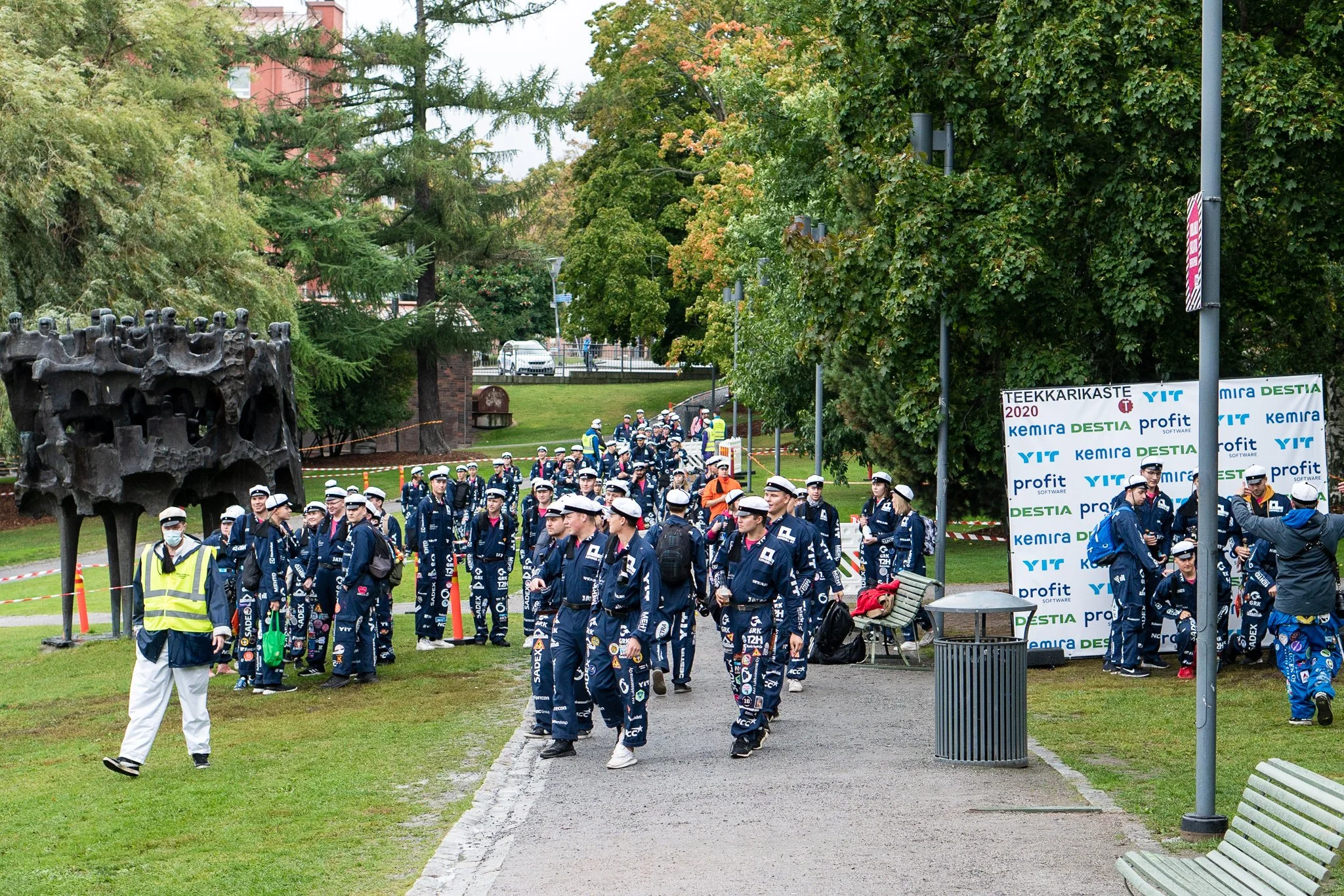 A group of people dressed in navy blue uniforms and white hats walking through a park, some wearing face masks. There are trees, benches, and a sculpture statue in the background, along with a sign and some fencing.