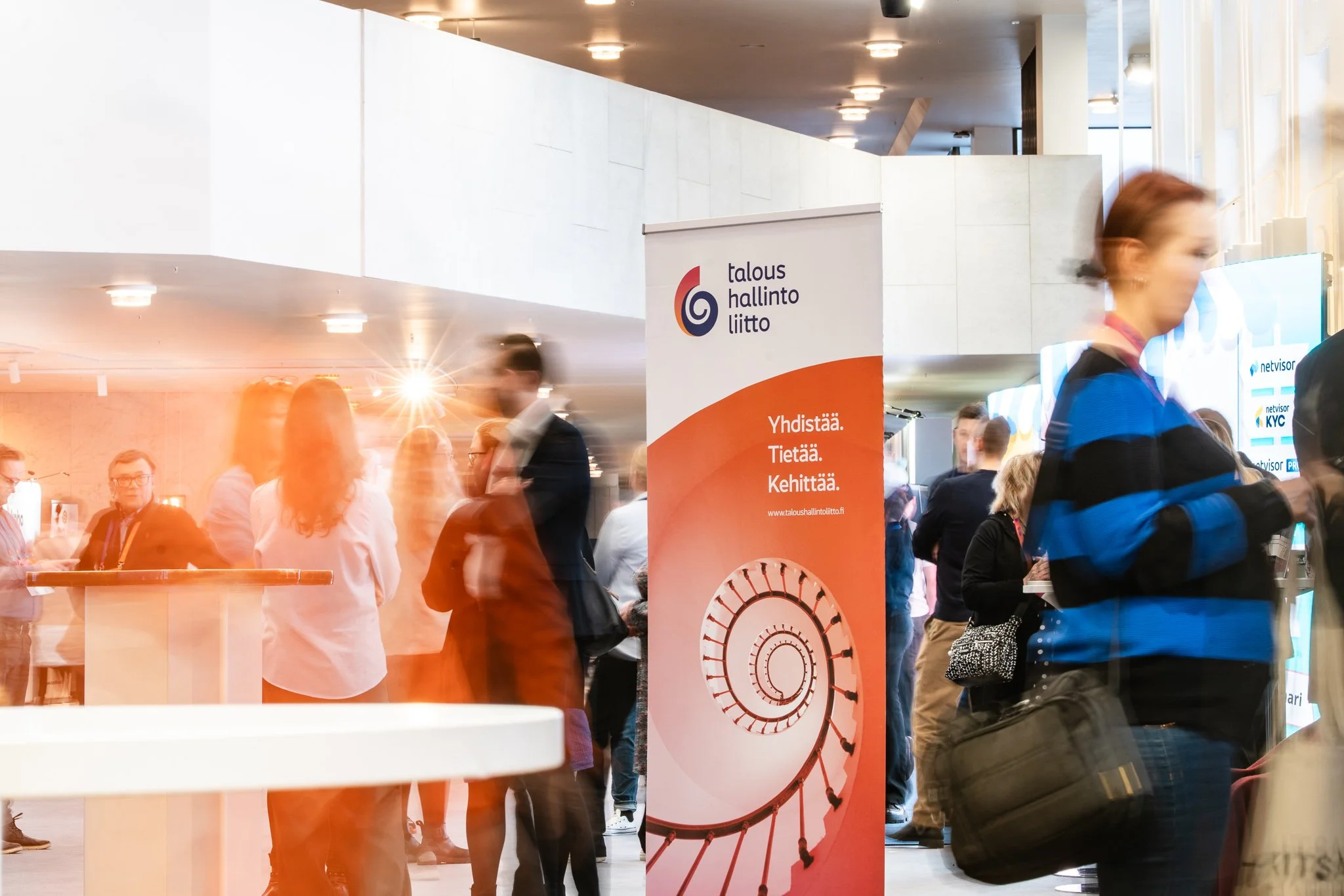 Conference or networking event with people standing and talking near a sign that reads 'Talous hallinto liitto' with a spiral graphic and Finnish text.