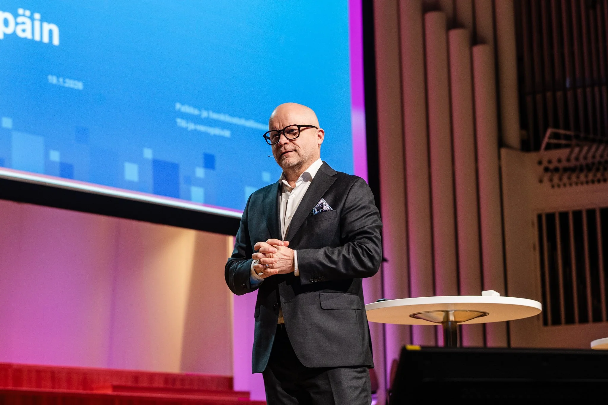 Bald man with glasses in a dark suit standing on stage with hands clasped, in front of a large screen with blue background and white text, in a conference or presentation setting.