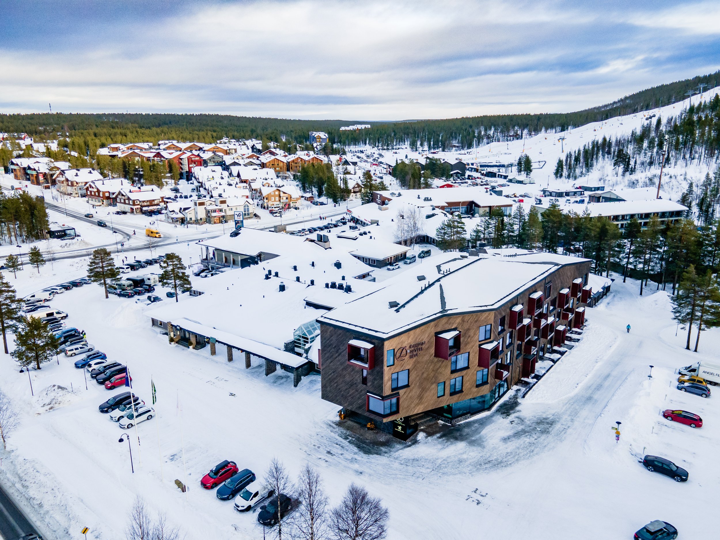 A snow-covered village with many houses and buildings surrounded by trees and mountains. A large modern hotel with red balconies and a parking lot in the front.