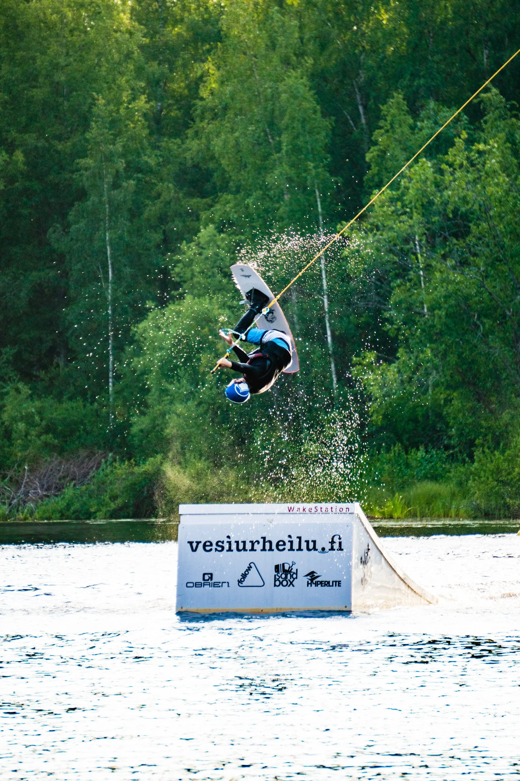 A person performing a wakeboarding trick on a ramp at a wakeboarding park, with a lake and dense green forest in the background.