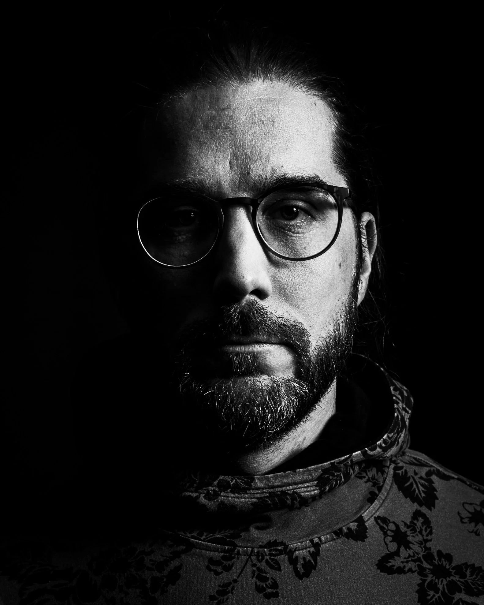 Black-and-white portrait of a man with glasses, beard, mustache, and long hair, wearing a patterned shirt, against a dark background.