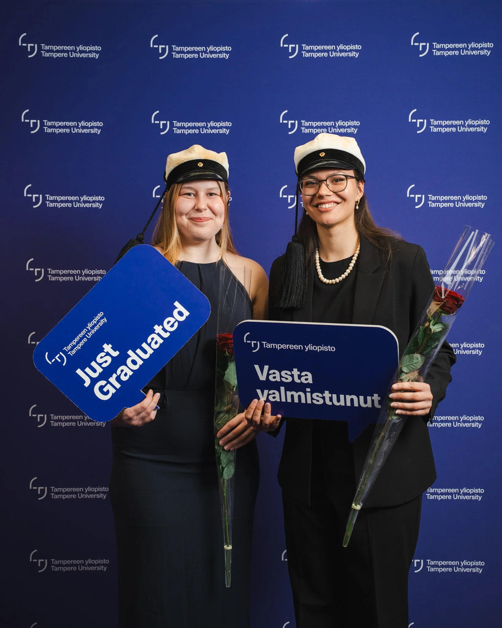 Two women wearing graduation caps and holding signs and roses, standing in front of a blue Tampere University backdrop, celebrating graduation.