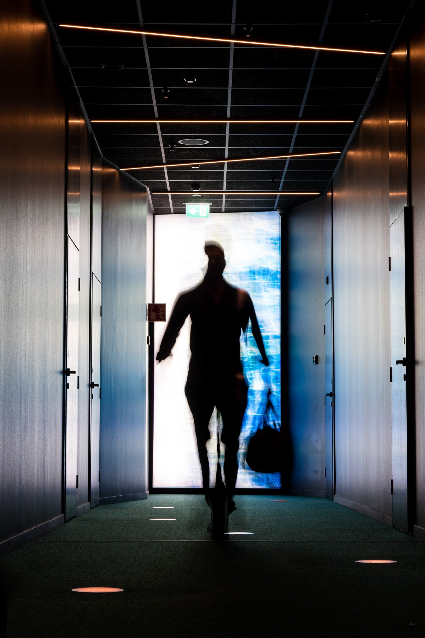 Silhouette of a person holding a bag walking through a modern hallway with illuminated ceiling and light spots on the floor, backlit by a large glass door or window.