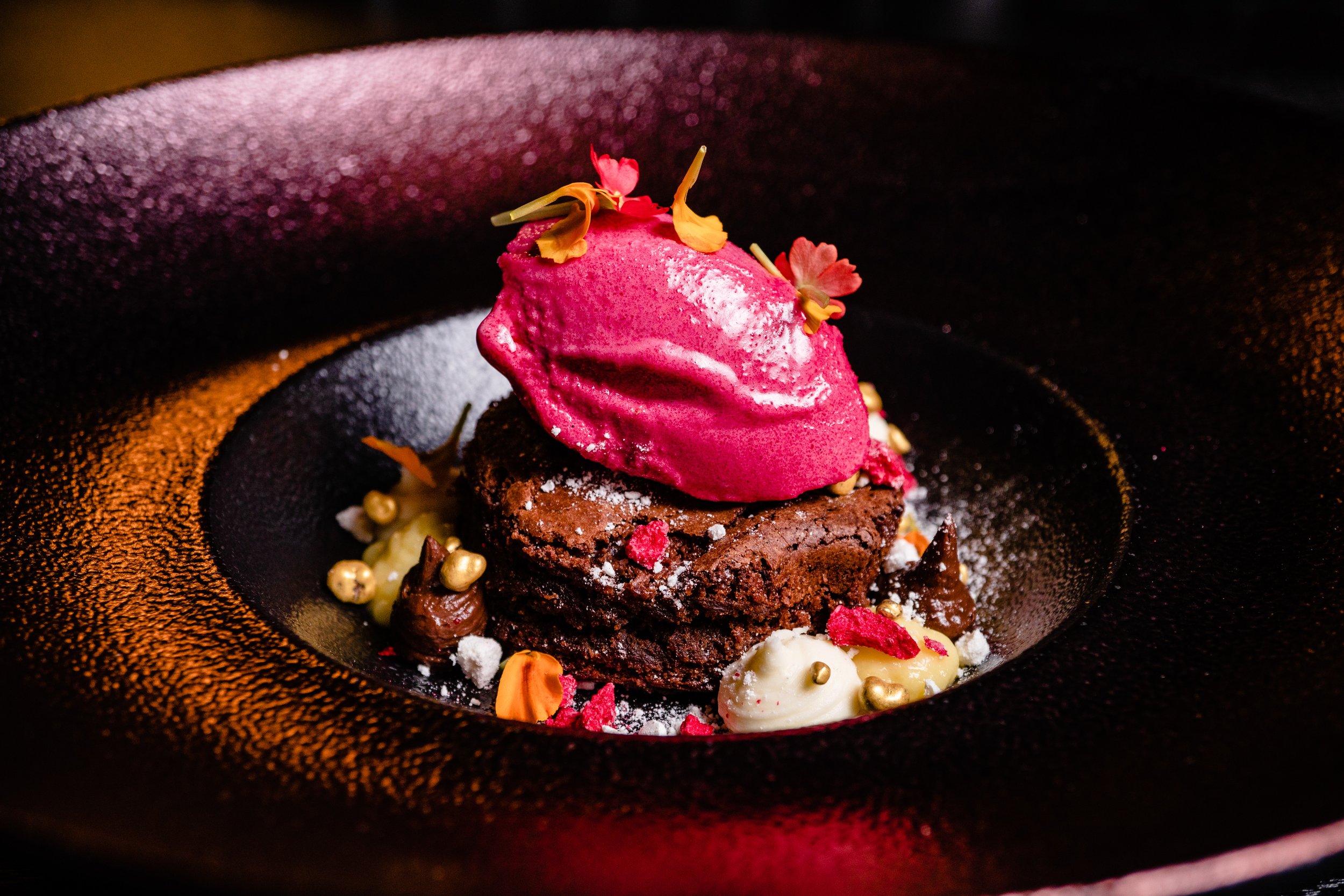 Chocolate dessert topped with a scoop of pink sorbet, edible flowers, and garnished with small decorative elements on a black plate.