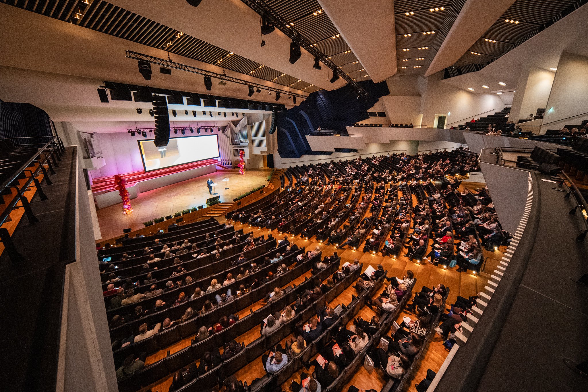 Large auditorium filled with seated audience watching a presentation on stage with large screen, decorated with balloons