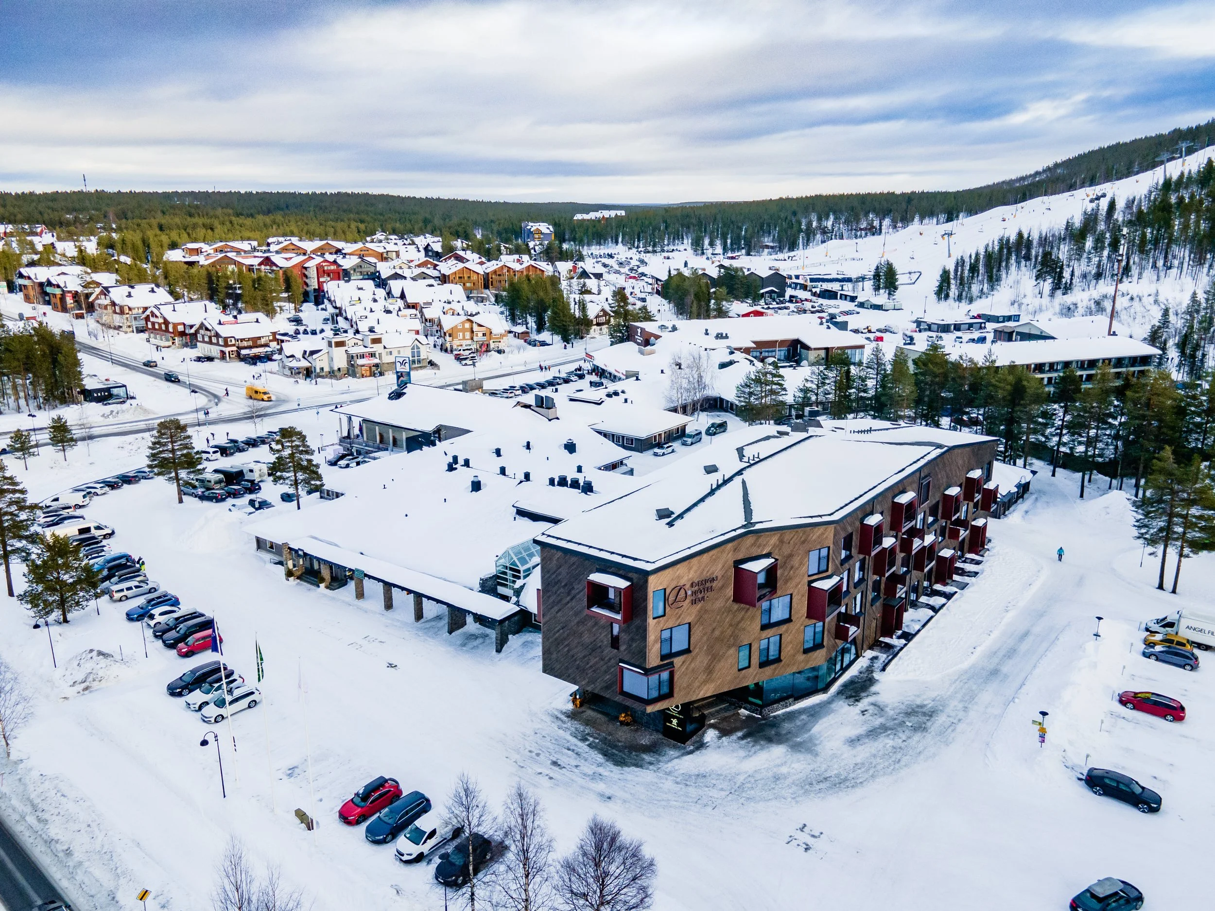 Aerial view of a snowy town with a mix of residential and commercial buildings, surrounded by snow-covered trees and hills, on a cloudy day.