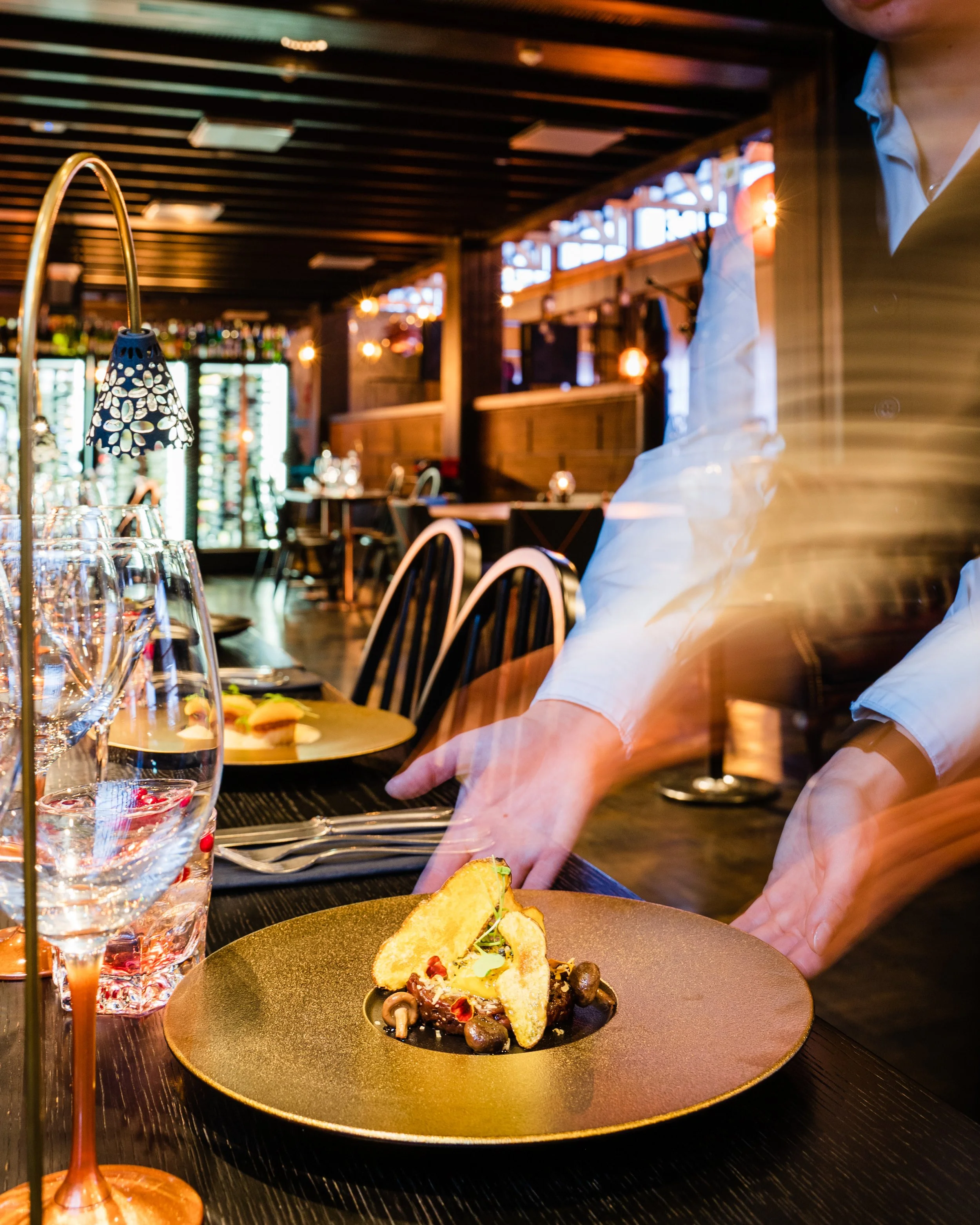 A restaurant dining setup with glassware, silverware, and a plated gourmet dish. A server is placing or adjusting the dish on the table. The background features a bar area with bottles and warm lighting.