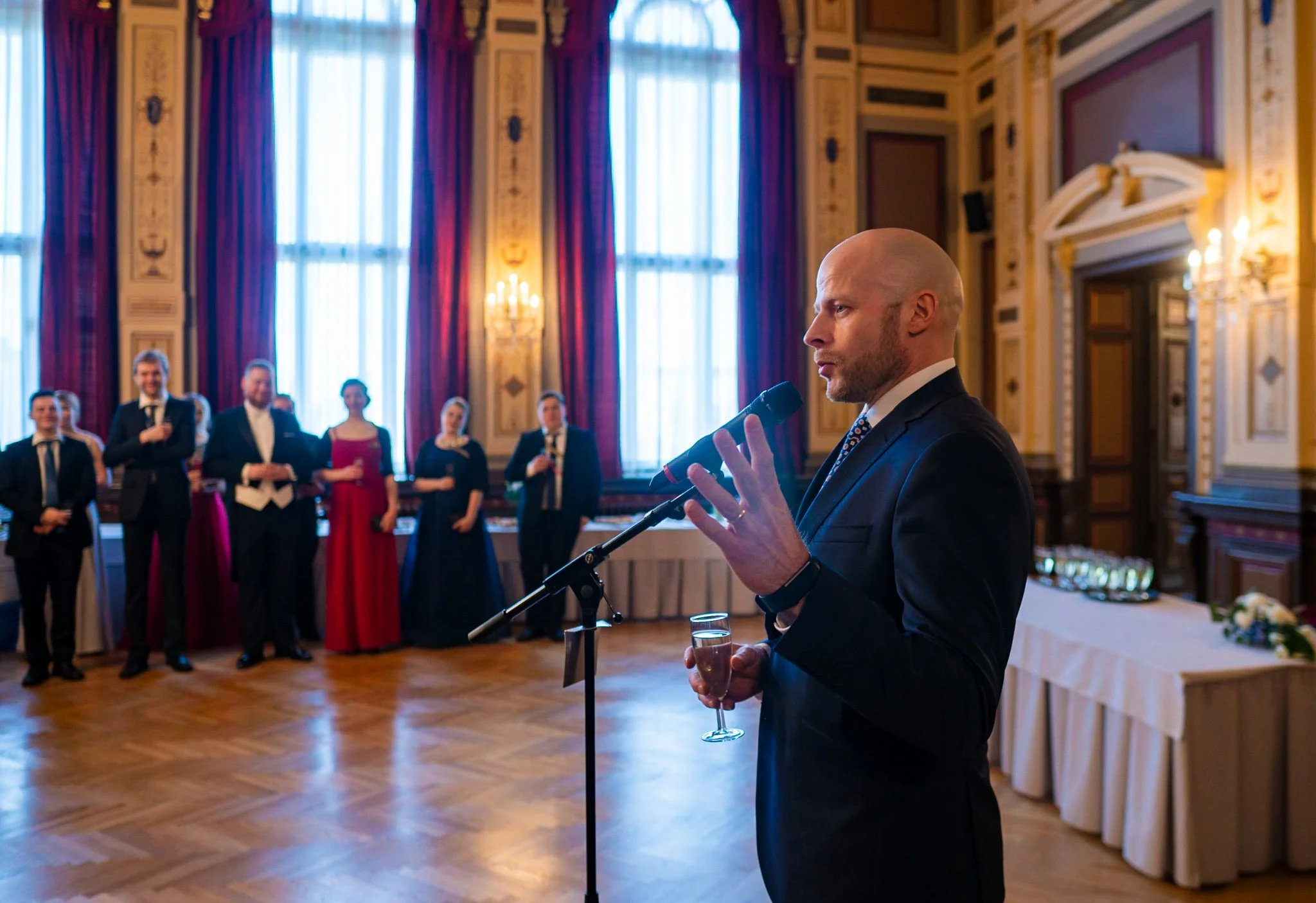 A man in a suit giving a speech at a wedding reception, with guests standing and listening in a grand, ornately decorated room with tall windows and drapes.