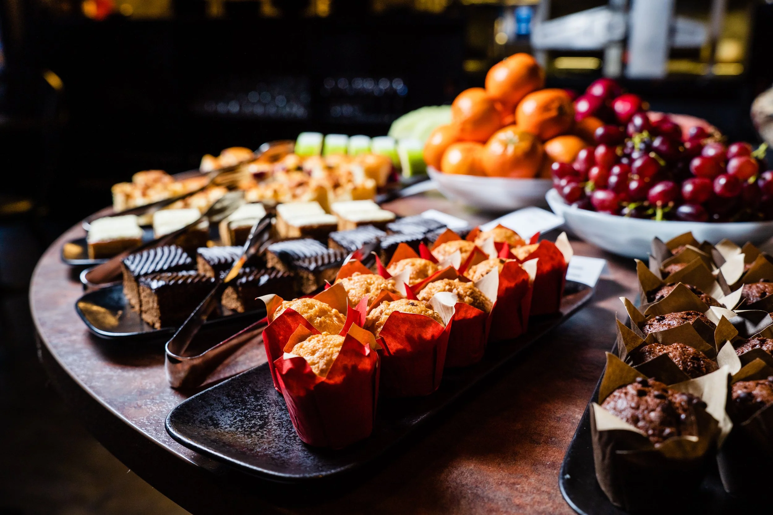 Assorted desserts including muffins, cakes, and chocolate treats on black trays, with bowls of grapes and tomatoes in the background.