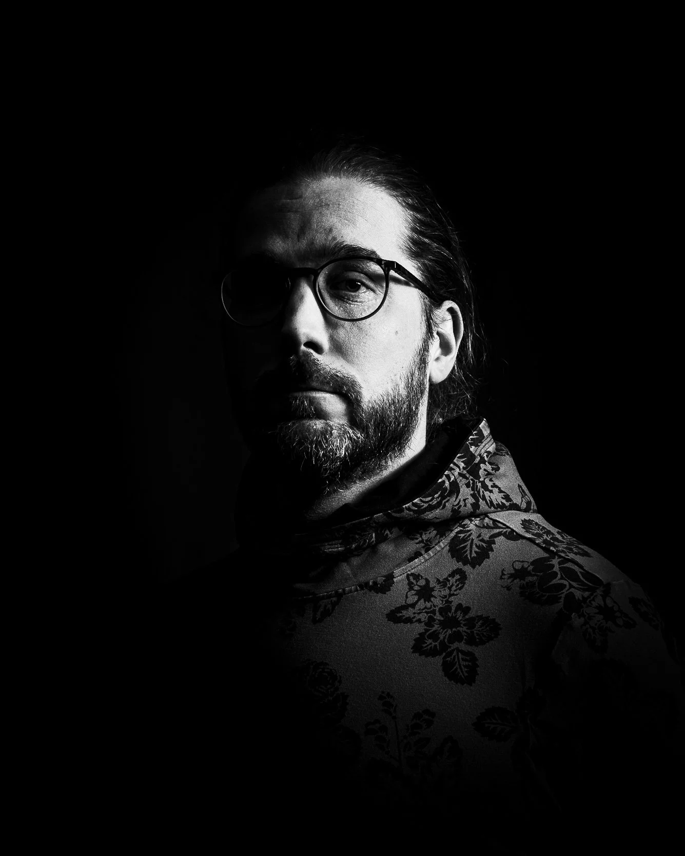 A black and white portrait of a man with glasses, a beard, and long hair, wearing a patterned shirt, with a dark background and dramatic lighting.