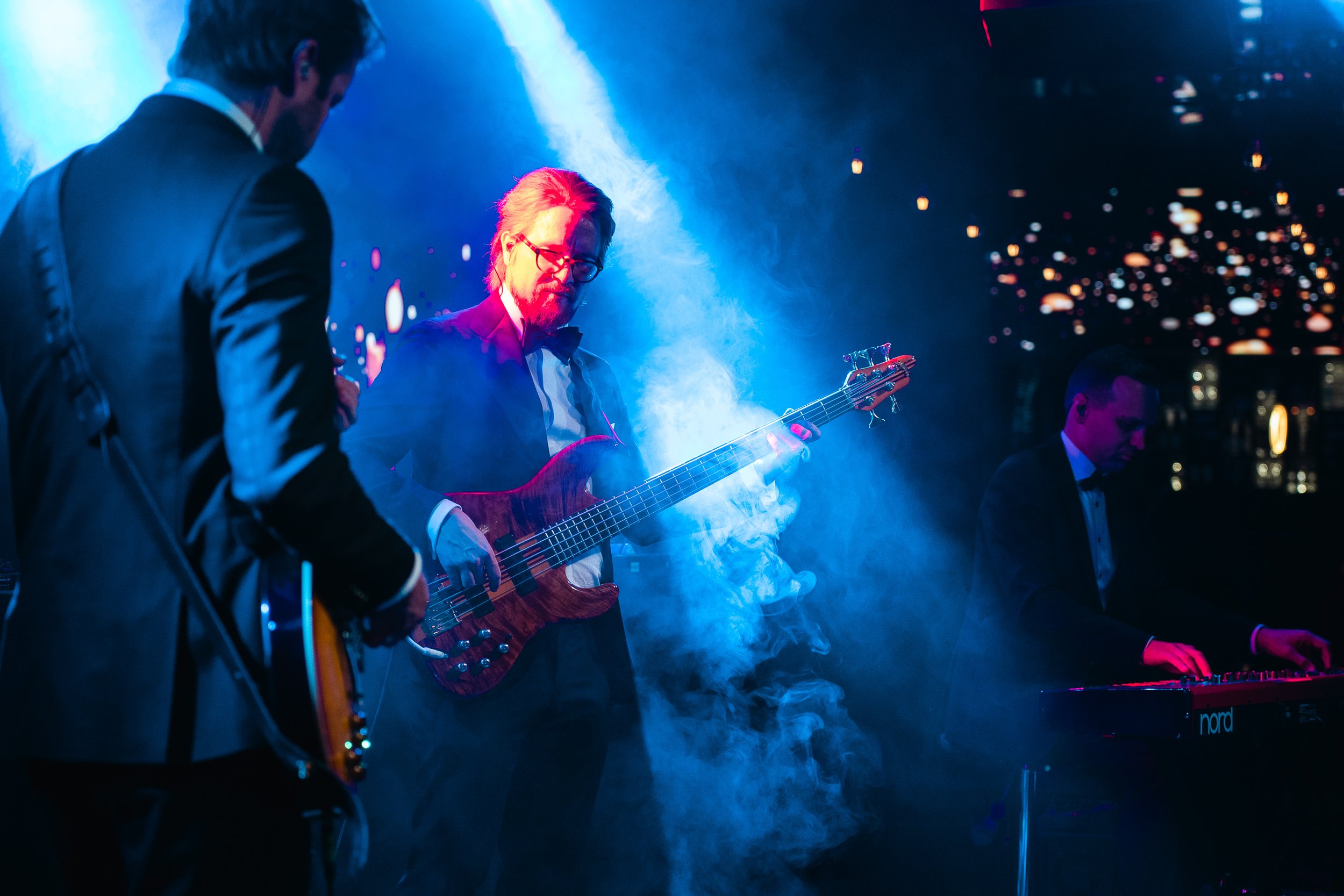 Three musicians performing on stage with colorful lighting and smoke, one playing electric guitar, one playing bass guitar, and one playing keyboard.