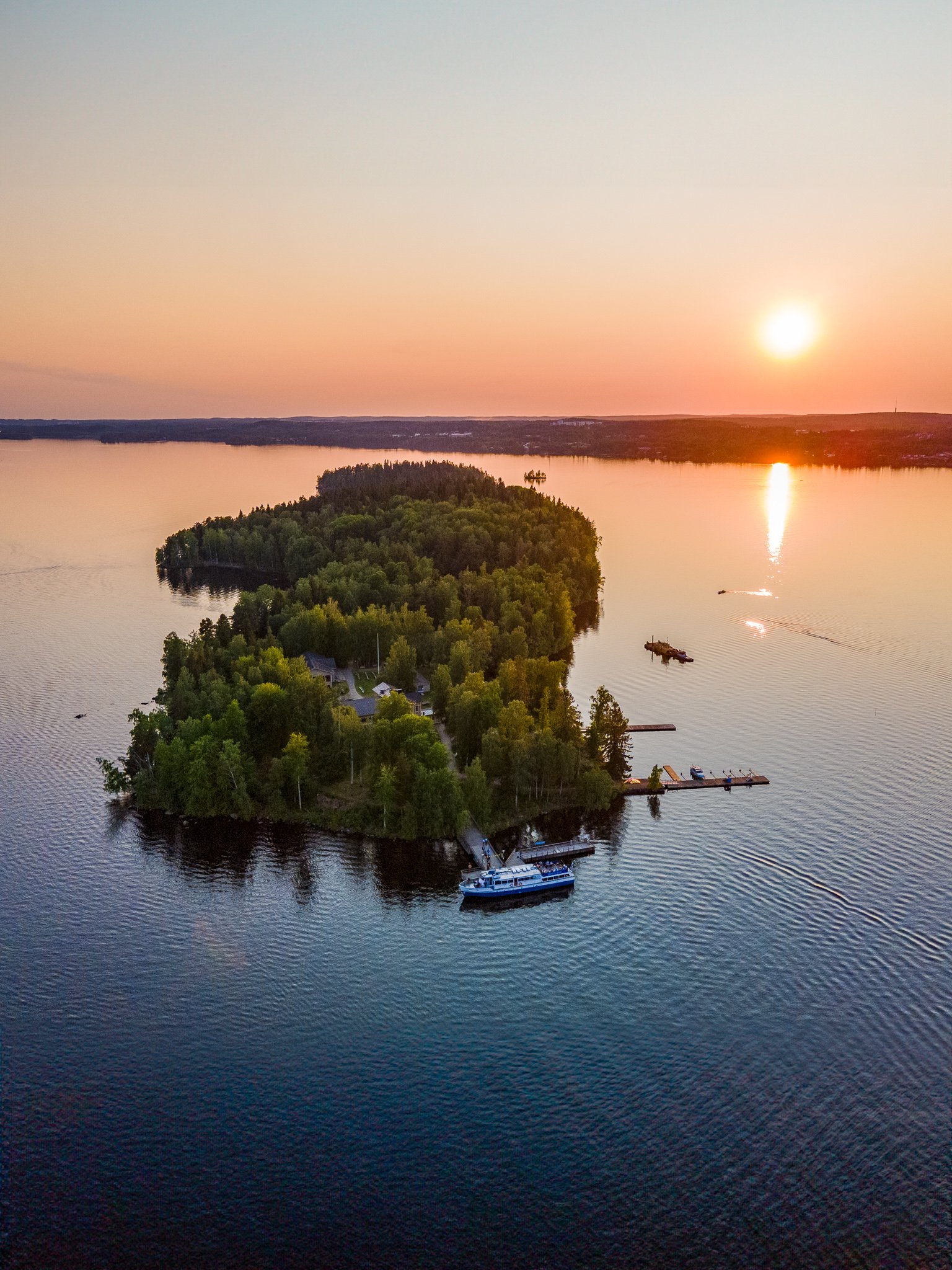 Aerial view of a small, forested island with a few buildings and docks, surrounded by a calm lake at sunset.