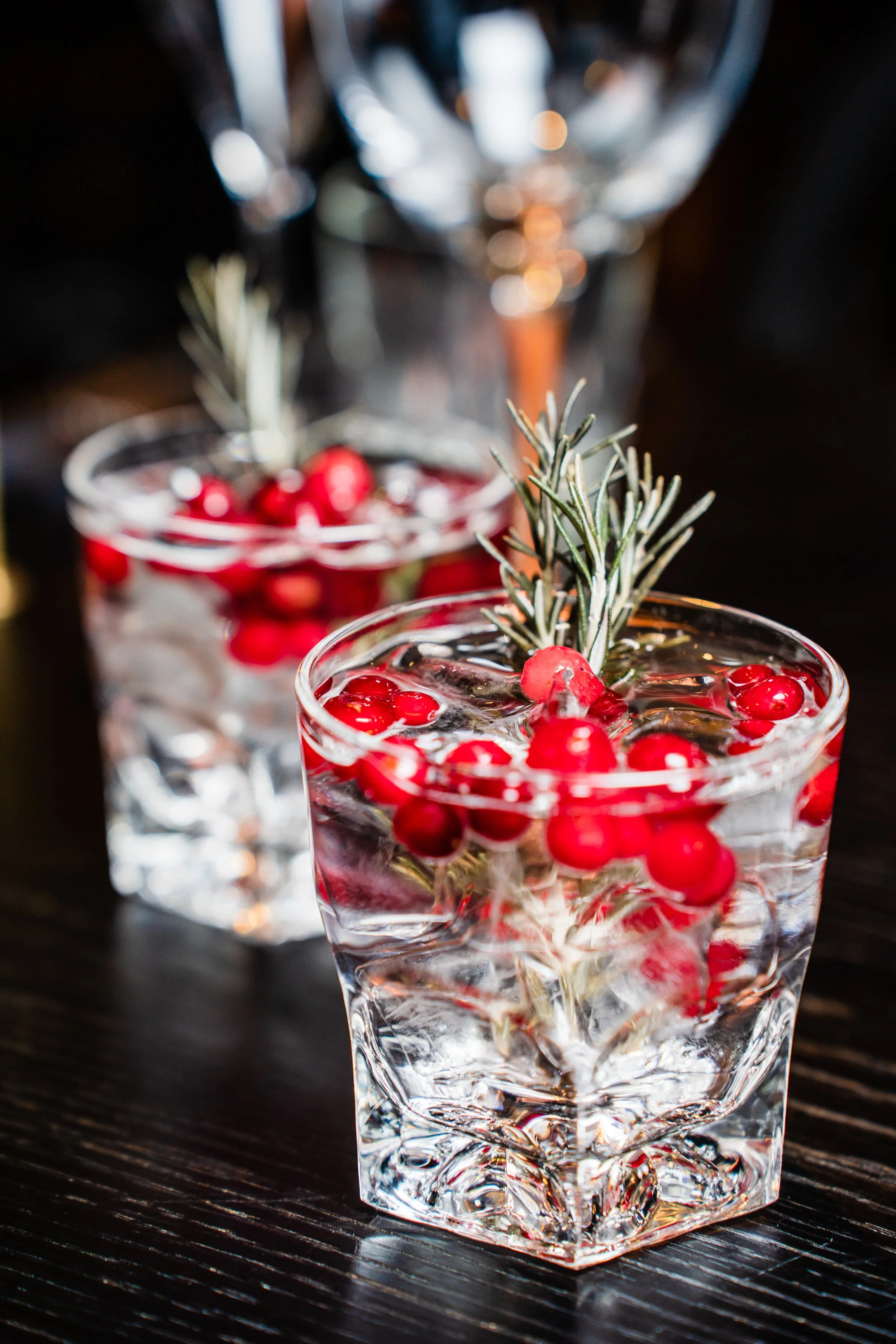 Two glasses of clear beverage garnished with red berries and rosemary sprigs on a dark wooden table