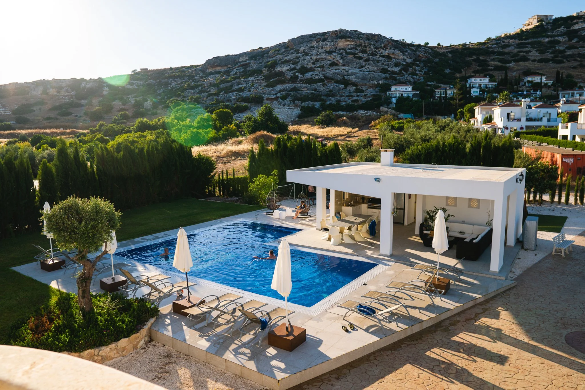 View of a modern house with a swimming pool, people swimming, surrounded by lounge chairs and umbrellas, with green trees and hillside in the background during daylight.