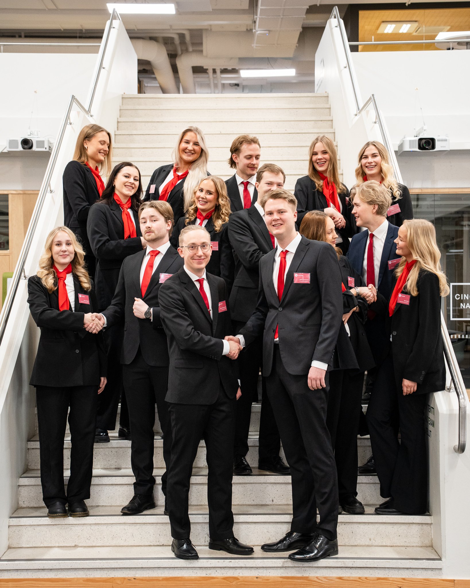 Group of young professionals in business attire, standing on stairs, shaking hands, smiling, in a modern office building.