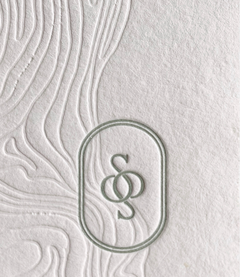 Close-up of textured white paper background with embossed design and a transparent oval outline containing the letters S and Q intertwined.