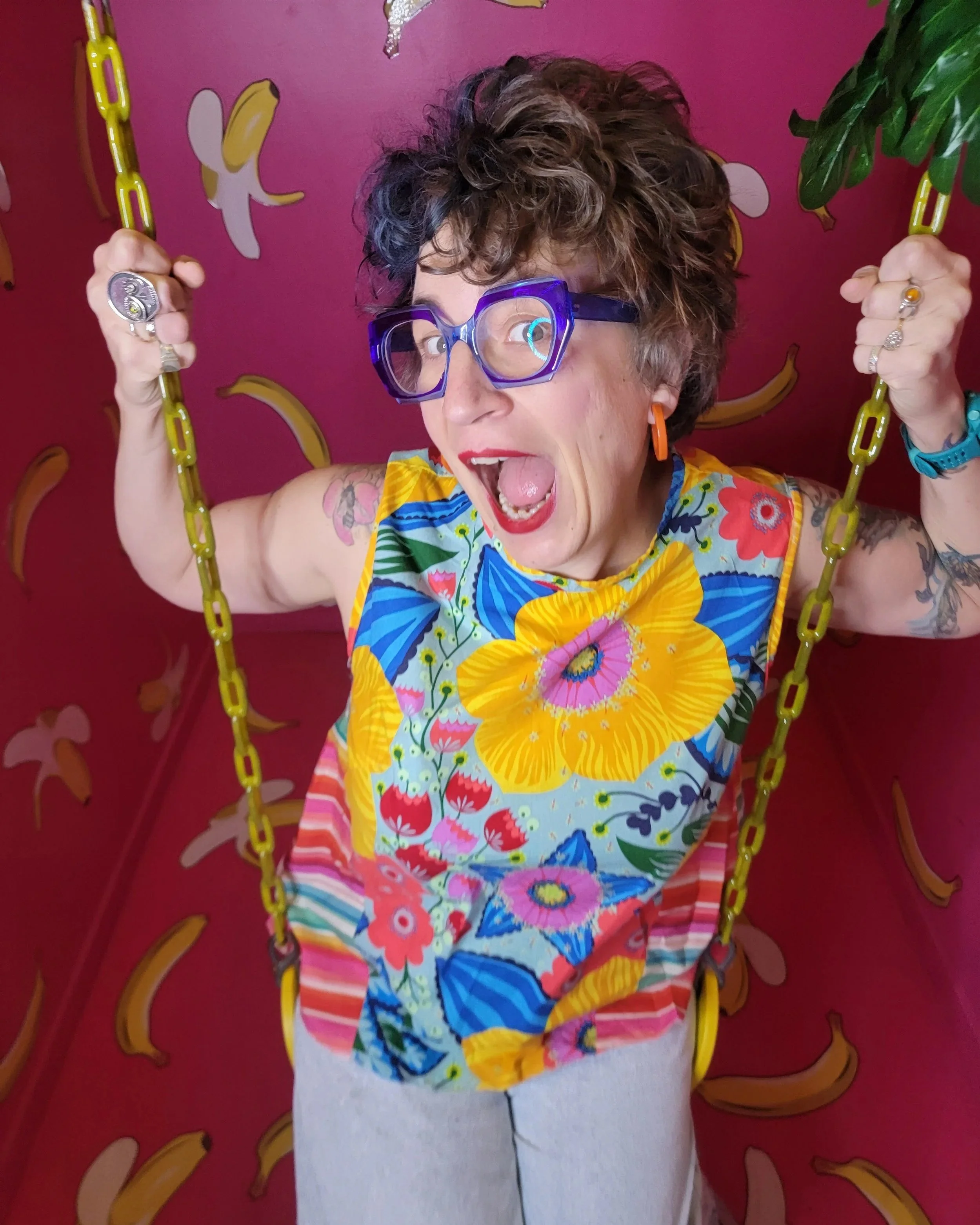 A woman with short, curly hair wearing large purple glasses, orange earrings, and a colorful floral top. She is sitting on a yellow swing with yellow chain links, against a pink background decorated with banana illustrations. The woman appears to be excited or surprised, with her mouth open and eyes wide.