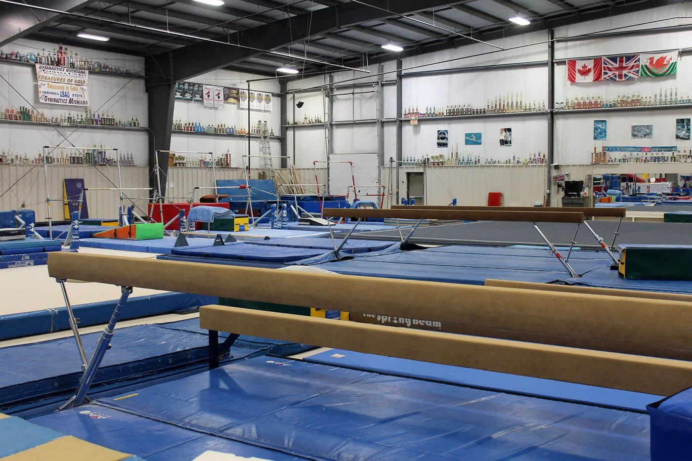 Indoor gymnastics training facility with various gymnastic equipment, including balance beams, mats, and uneven bars, decorated with flags and trophies displayed on shelves along the walls.