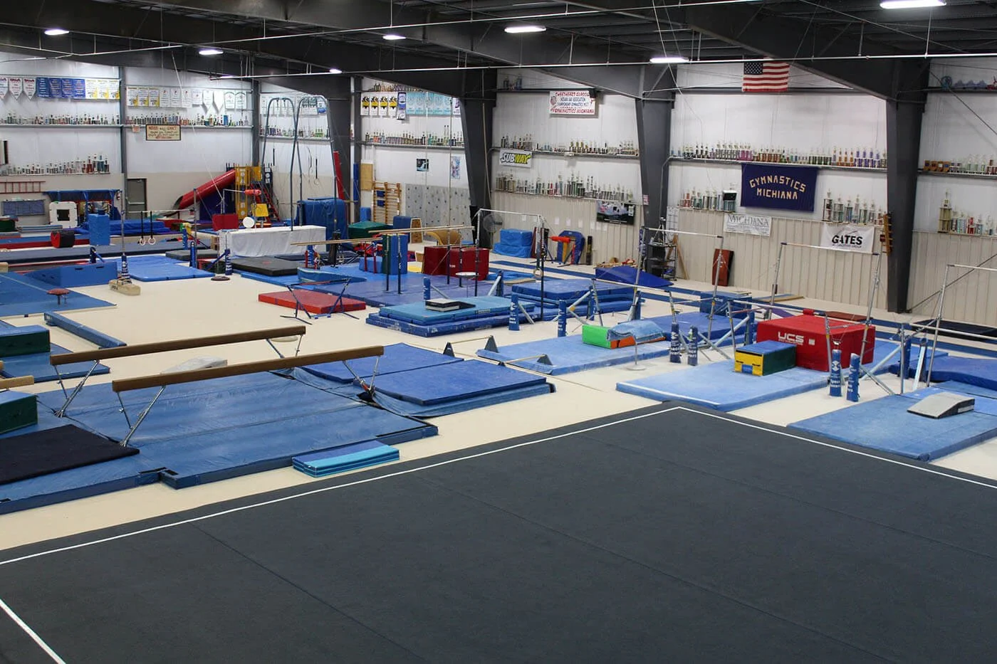 Indoor gymnastic training facility with mats, uneven bars, balance beams, vault and other gymnastics equipment, with banners and trophies on the walls.
