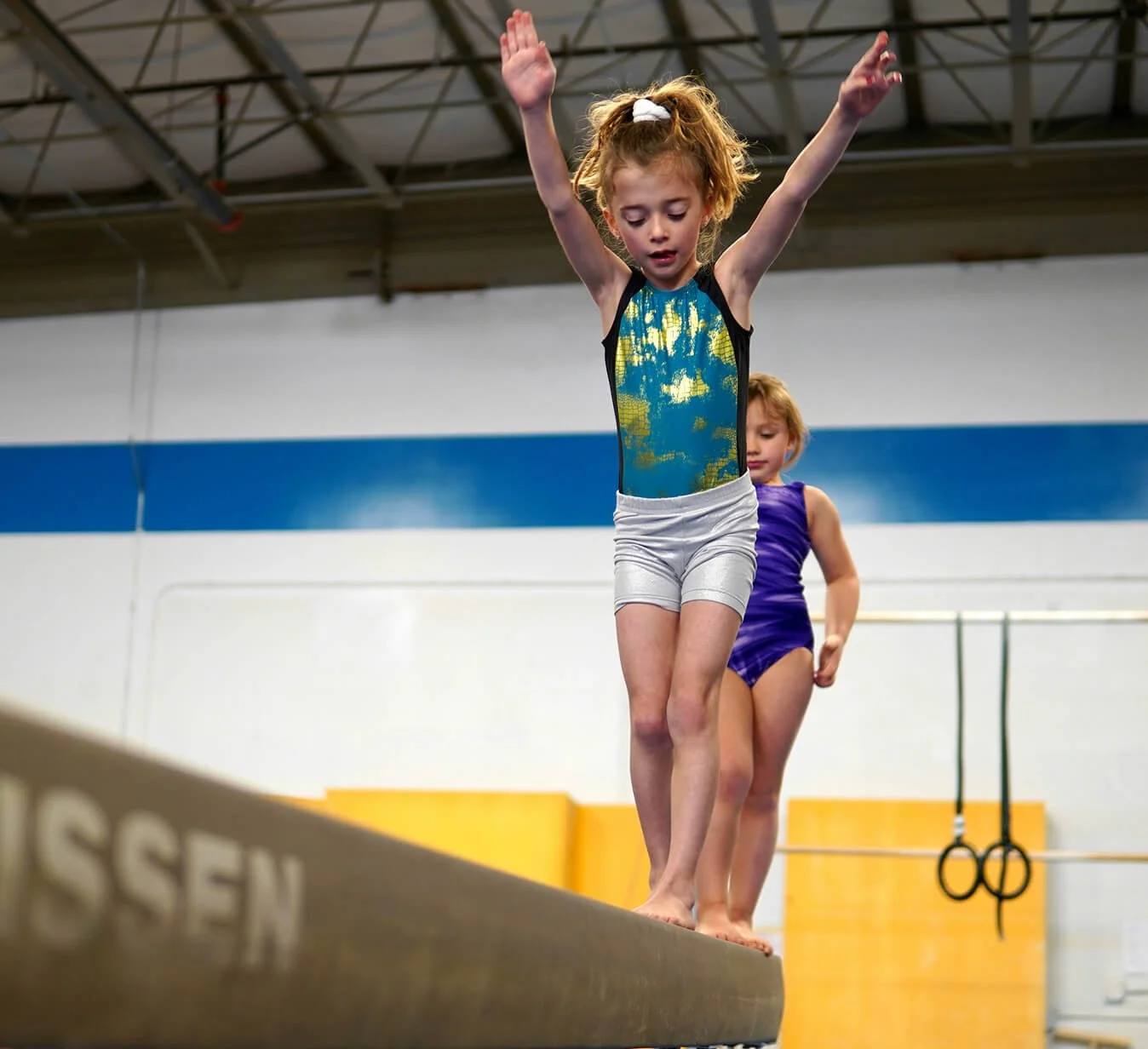 Young girl balancing on a gymnastics beam with arms raised, another girl standing behind her in a purple leotard, gymnasium background with rings and yellow padding.