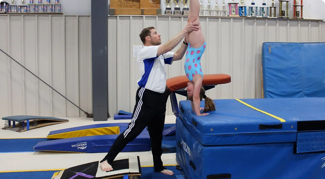 A young girl is performing a handstand on a gymnastics mat while an adult spotter supports her legs in a gym.