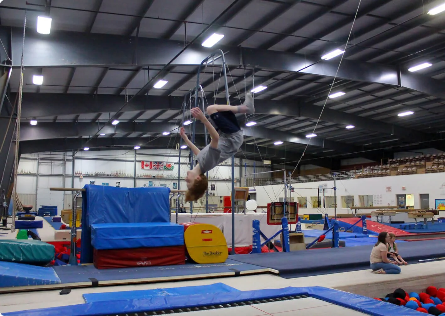 A gymnast performing on the uneven bars inside a gymnastics training facility, with safety mats and equipment around, while two people sit on the floor watching.