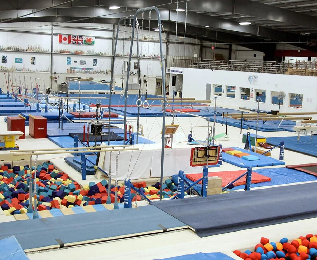 Indoor gymnastics facility with various equipment including trampolines, foam pits, balance beams, and rings, set up for training or competitions.