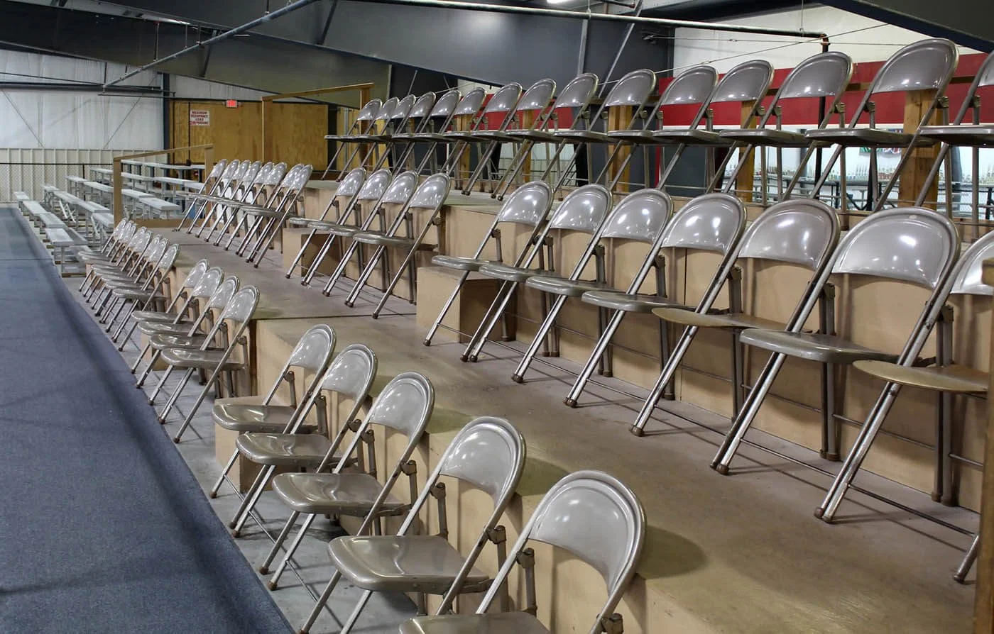 Multiple metallic folding chairs arranged in rows, positioned on a tiered wooden platform inside a sports stadium or arena.
