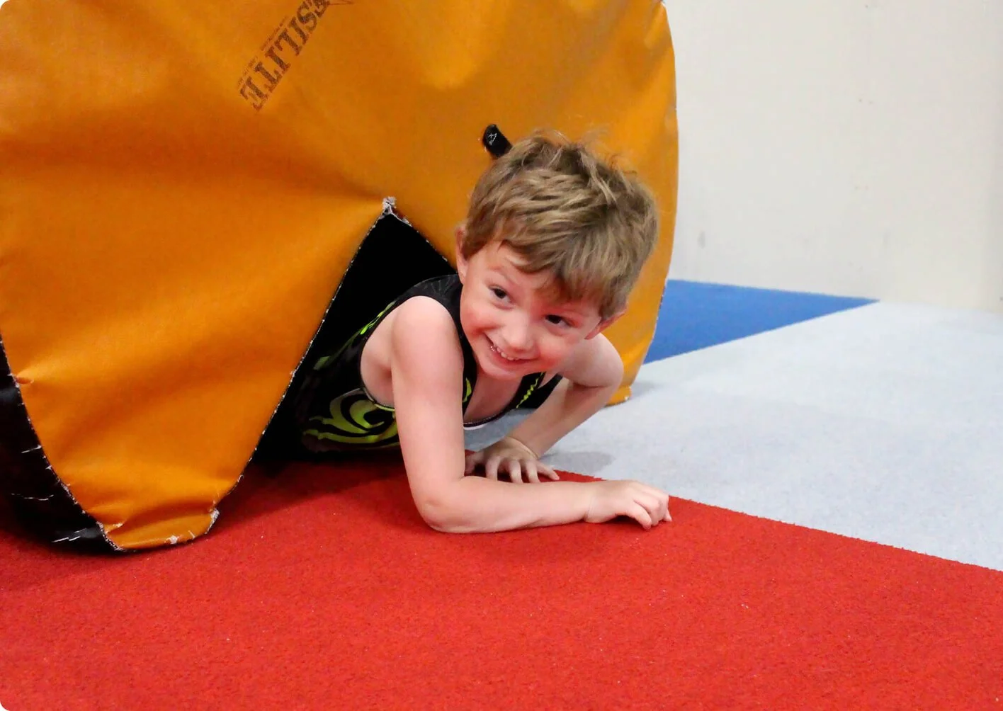 Young boy with light brown hair crawling through a black and yellow play tunnel on a red, gray, and blue gym mat.