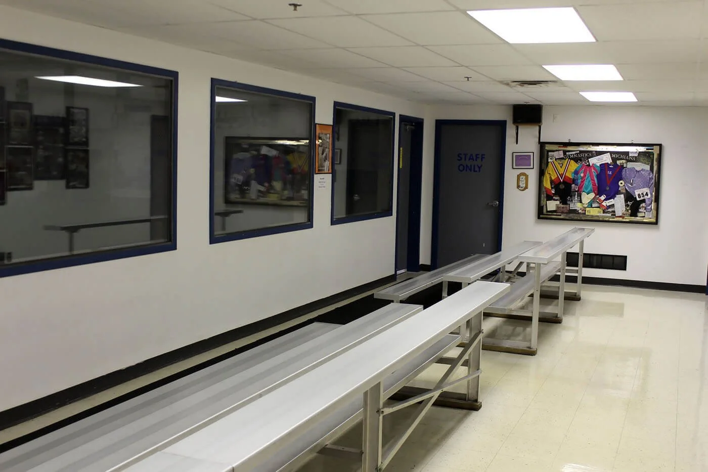 Empty waiting area with three metal benches, a door labeled 'Staff Only', and a bulletin board with gymnastics medals and photos.
