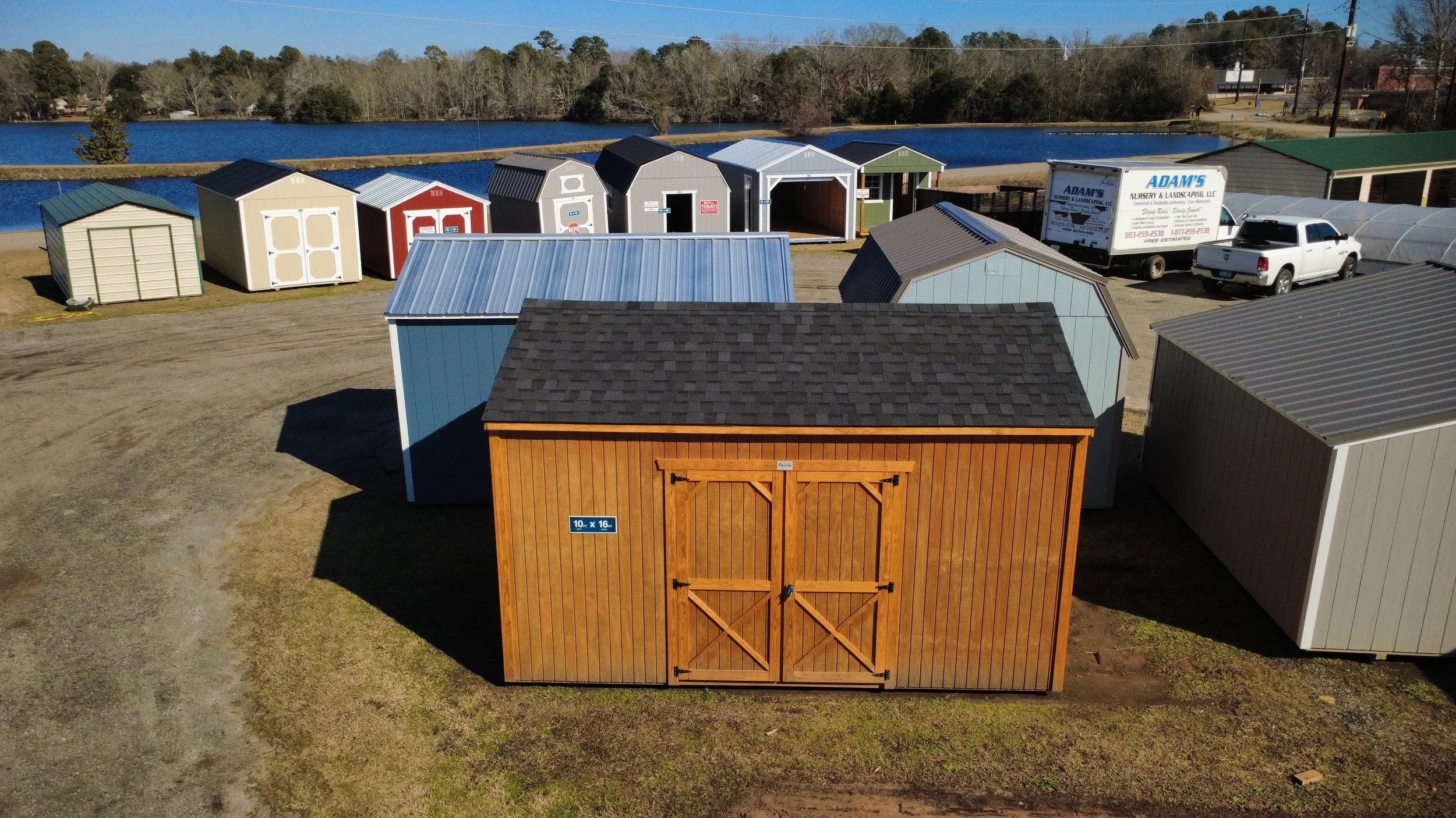Aerial view of storage sheds in various colors and sizes, with a pond and trees in the background. There are also trucks and a white pickup truck parked nearby.