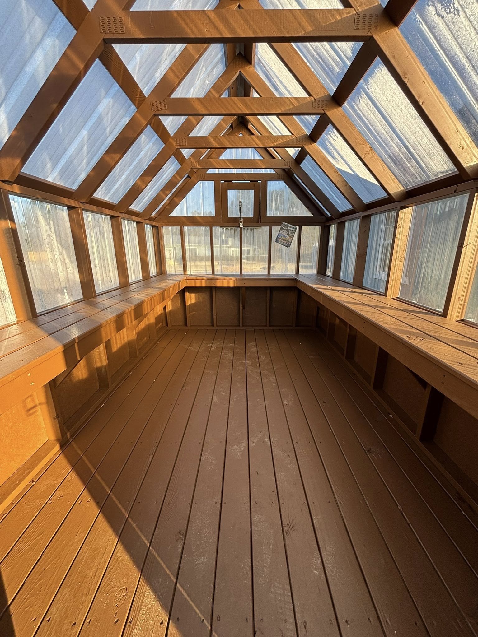 Interior of a wooden greenhouse with glass panels, wooden floor, and built-in benches along the sides. Sunlight is shining inside.