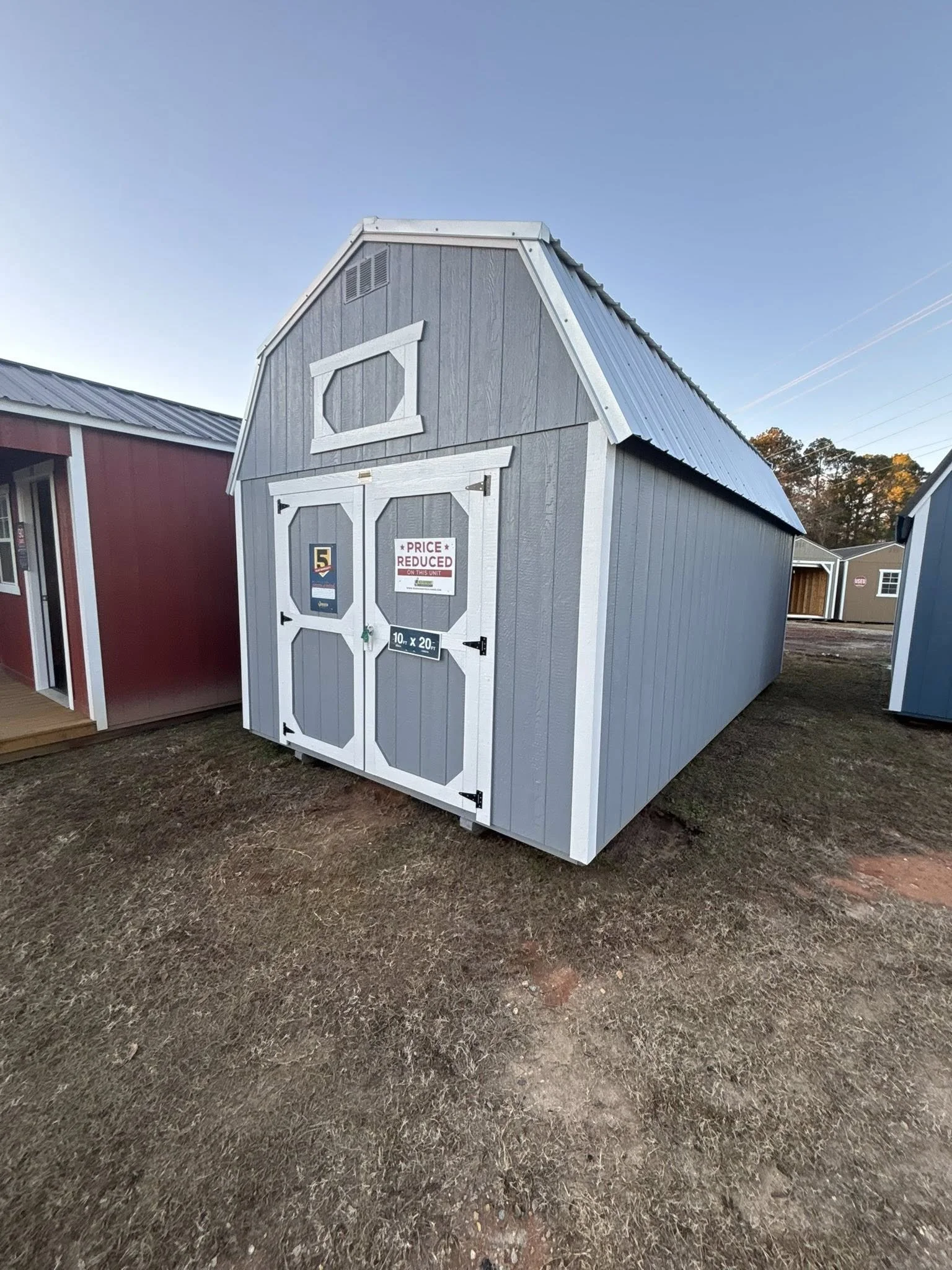 Gray and white barn-style shed with a double door, one window, and signs indicating a sale and size, situated on a dirt lot under a clear blue sky.
