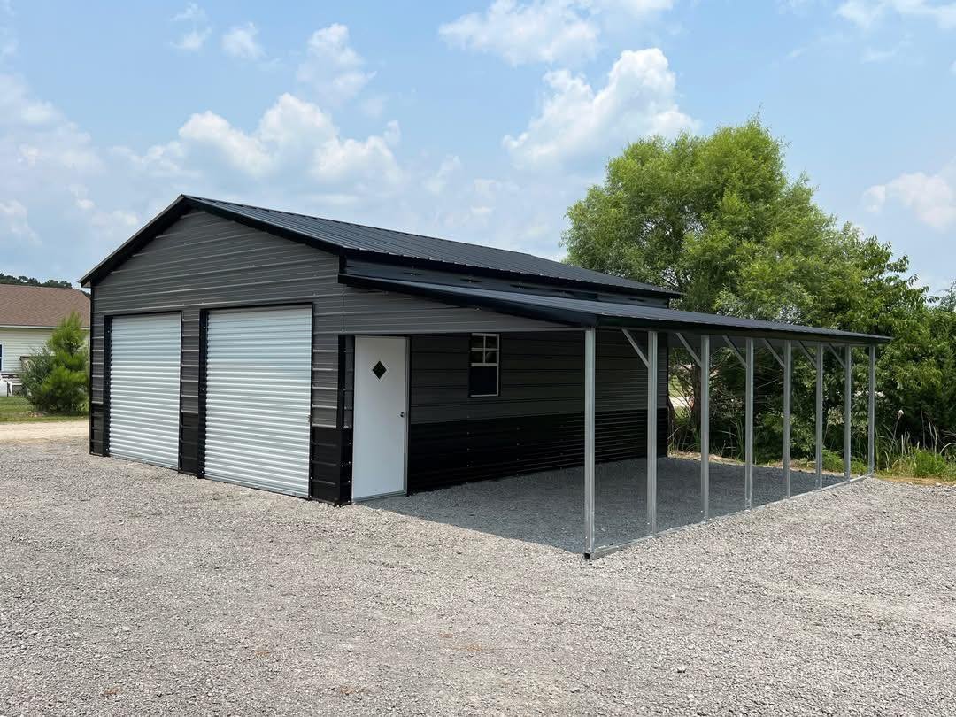 A black metal shed with a roof, featuring two large roller doors, a side door with a diamond-shaped window, a small window, and a metal carport extension on the right side, situated on a gravel lot with trees and residential houses in the background.