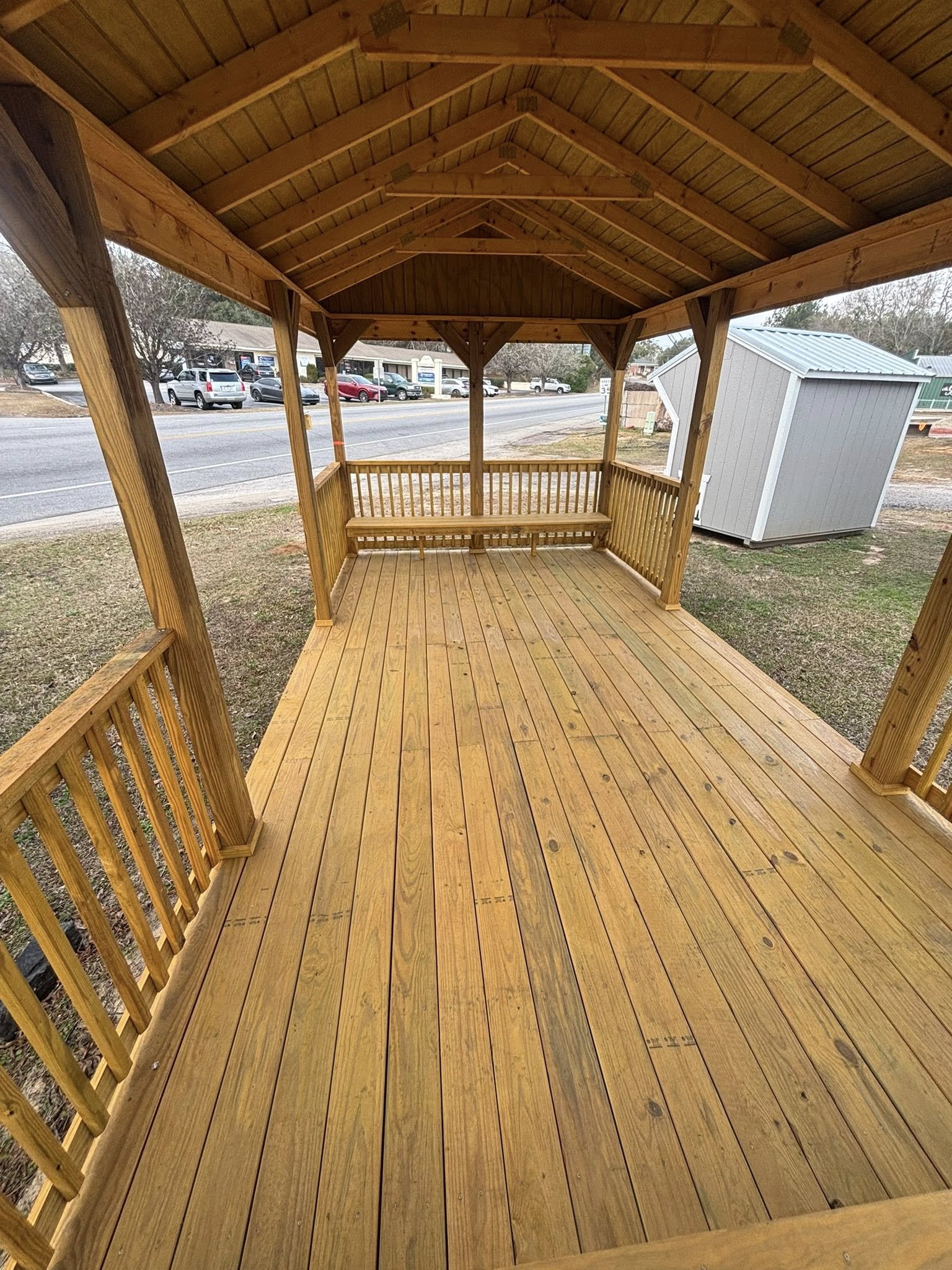 A newly constructed wooden gazebo with a polished wood deck and open sides, situated by a roadside with parked cars and a small gray shed nearby.